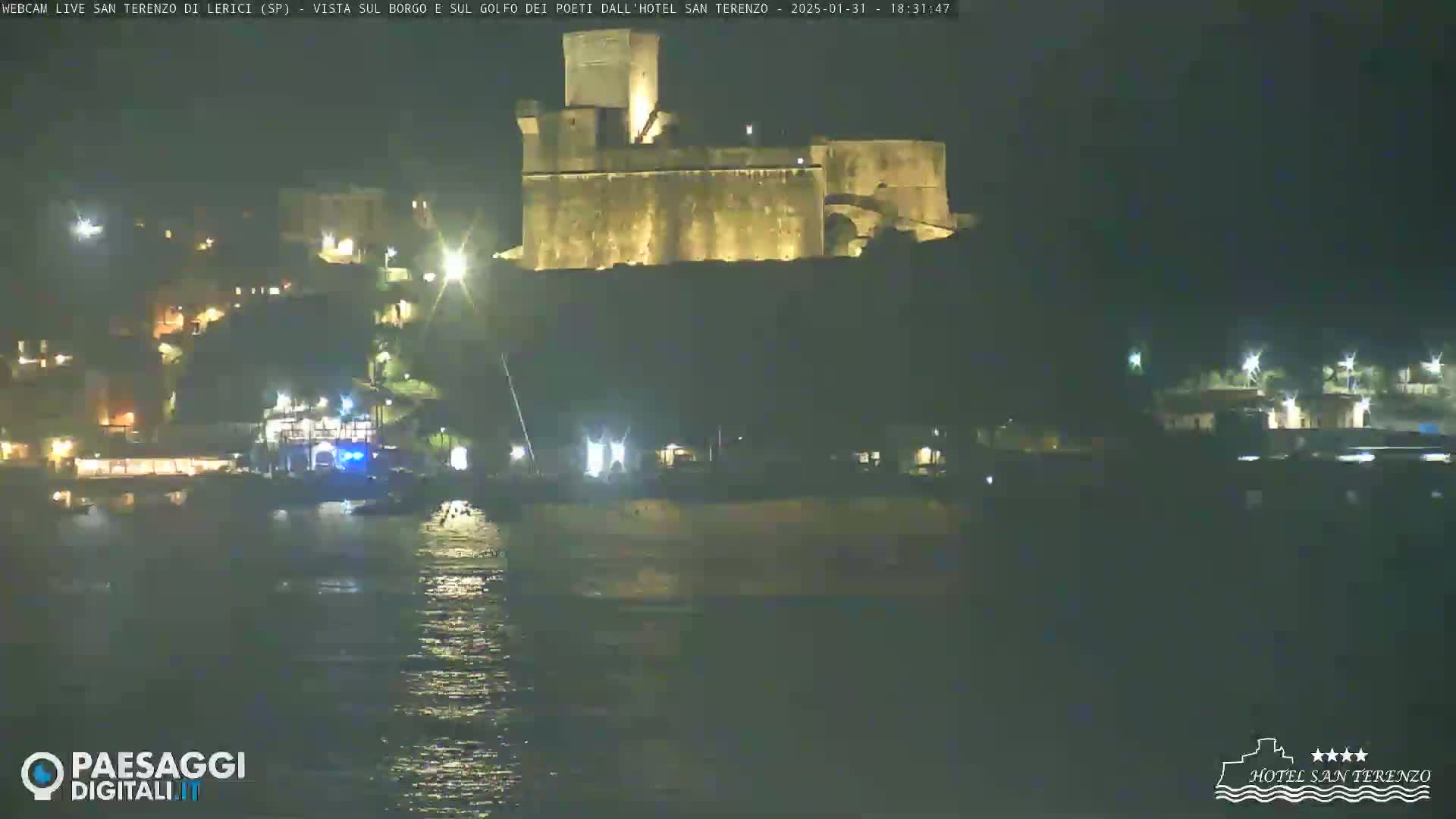 A nighttime view of a coastal town with a large illuminated castle overlooking a dark body of water.