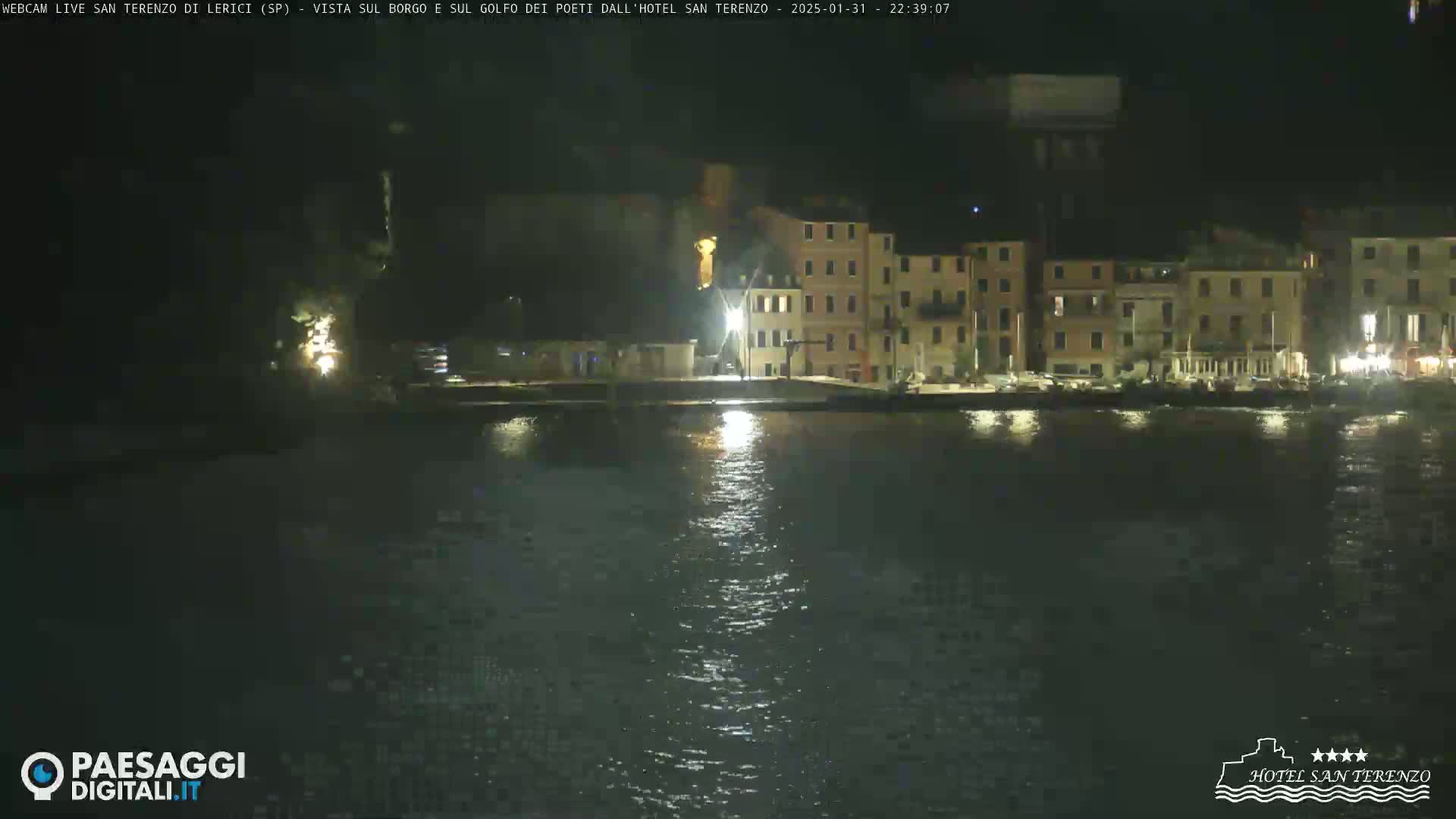 A nighttime view across calm water shows a row of illuminated buildings along a waterfront.