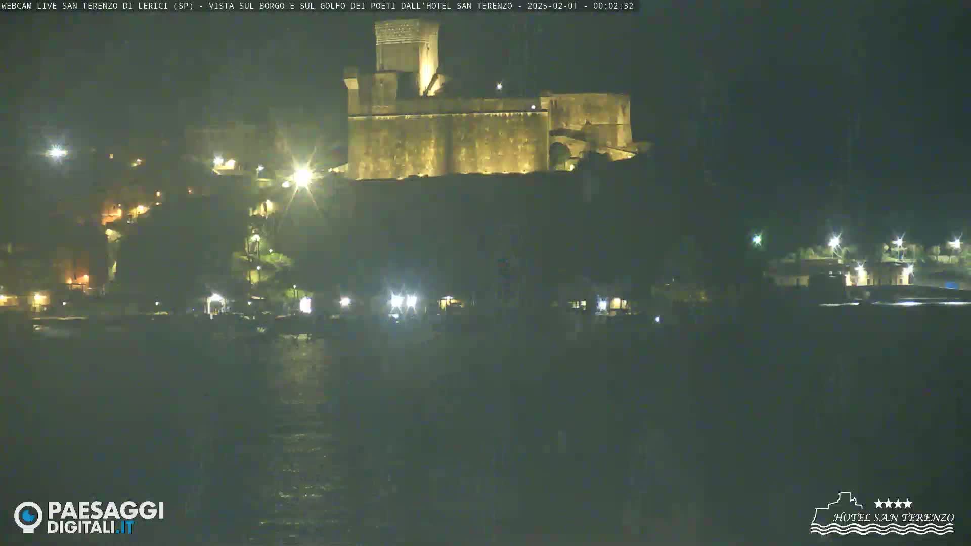 A nighttime view of a coastal town shows a large stone structure, possibly a fortress, illuminated against a dark, calm body of water.