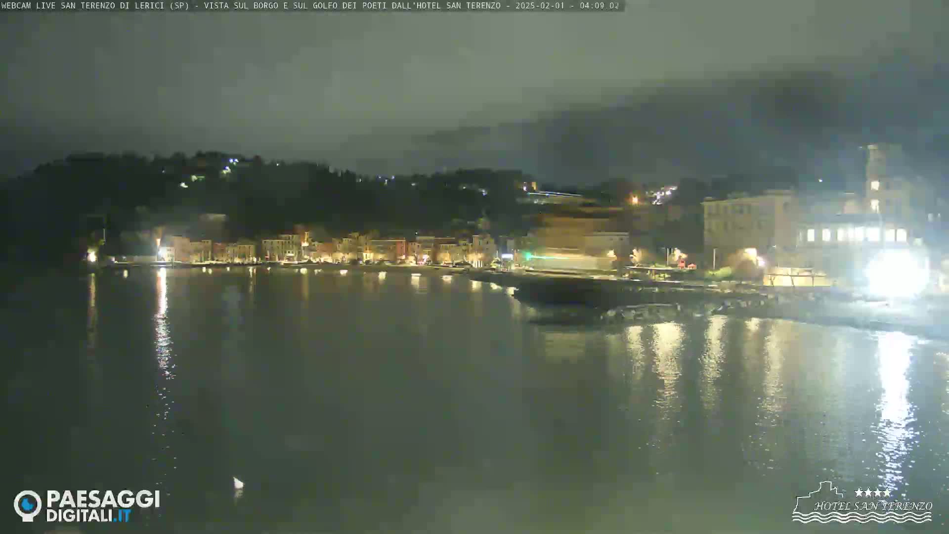 A nighttime view of a calm body of water reflecting the lights of a town nestled against a hill.