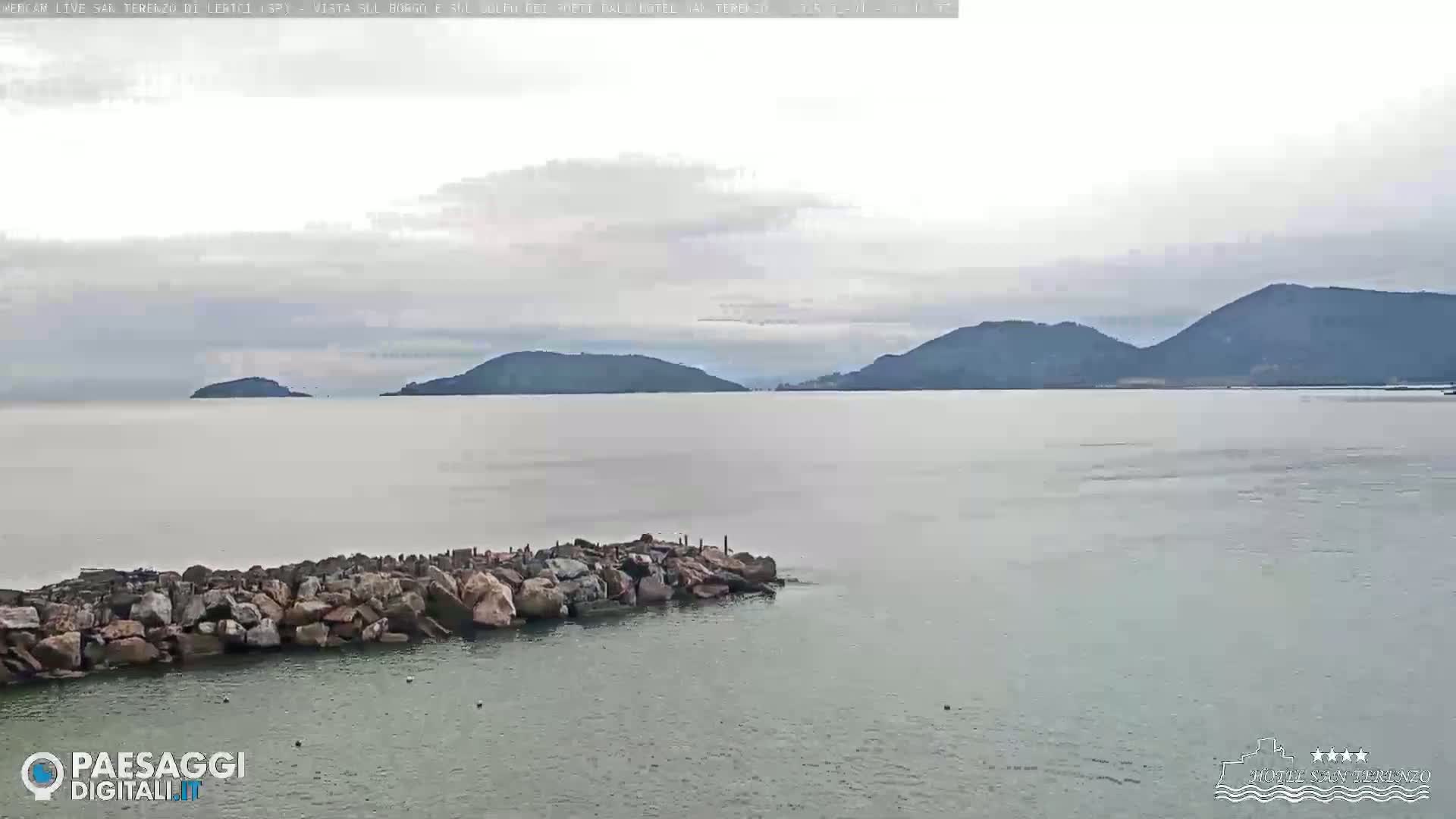 A rocky breakwater extends into a calm body of water under an overcast sky, with distant hills visible on the horizon.