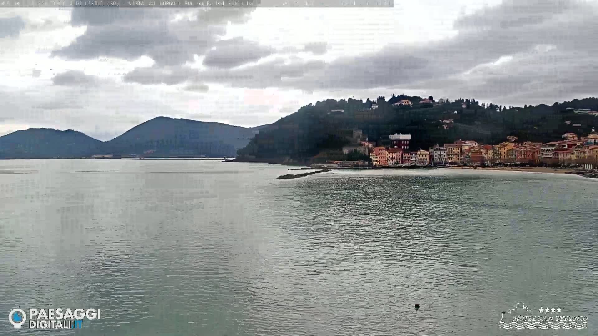 A calm body of water stretches towards a coastline where a town nestled against a hillside under a cloudy sky.