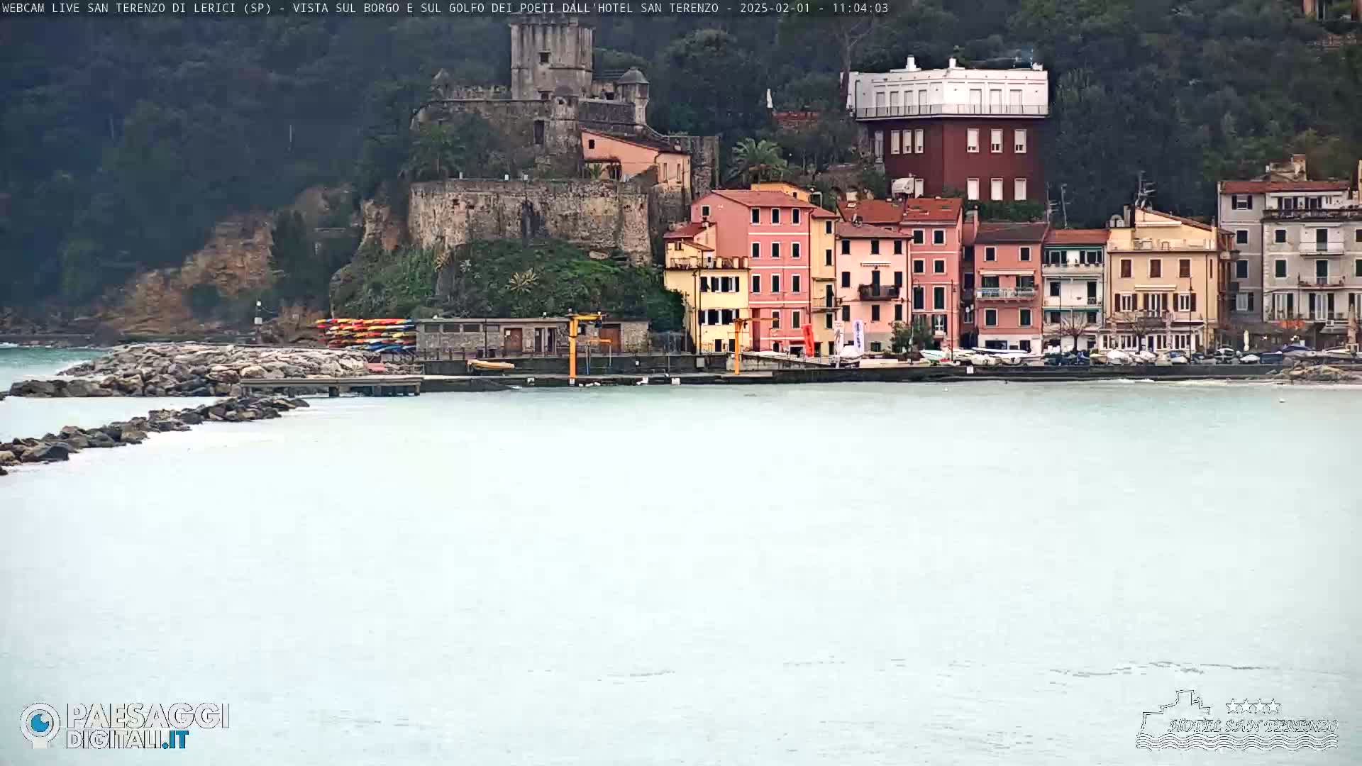 A calm, light-grey body of water sits in front of a collection of colorful buildings nestled against a hillside, with a stone fortress visible further up the hill.