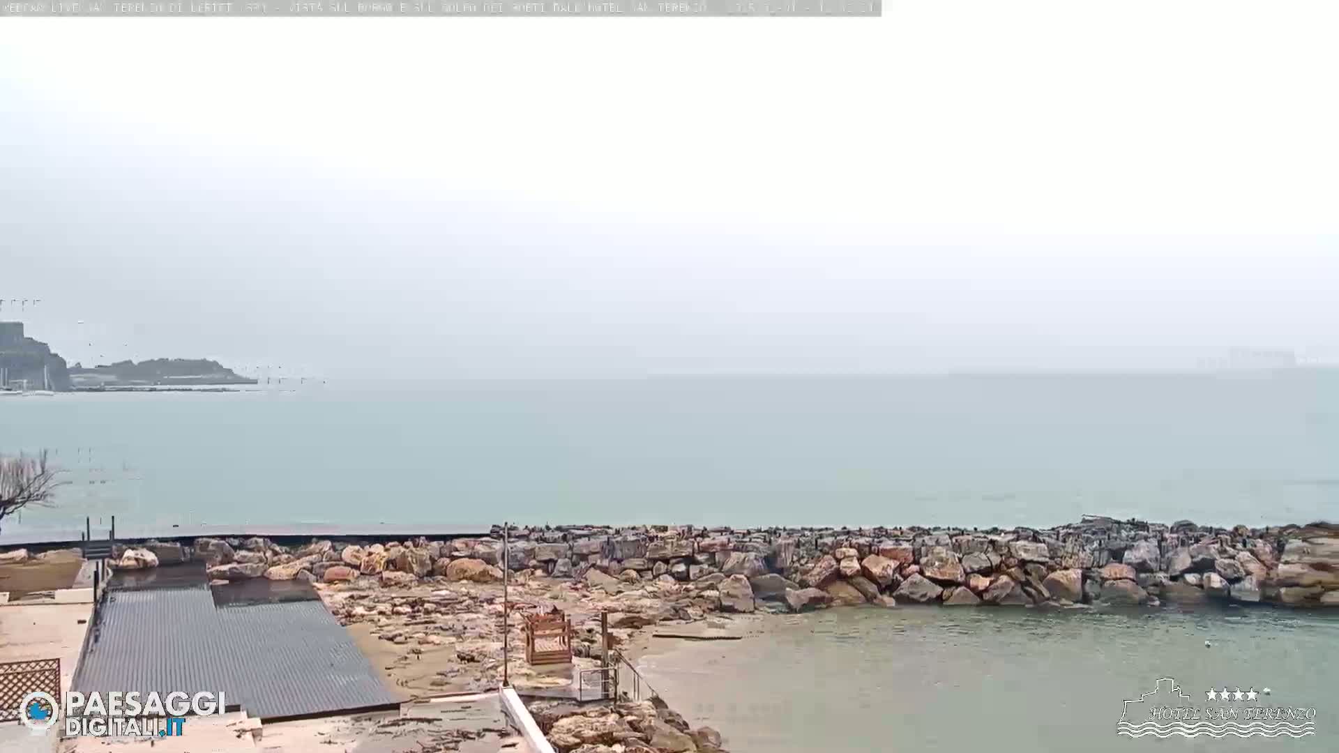 A rocky shoreline meets a calm, hazy sea under an overcast sky.