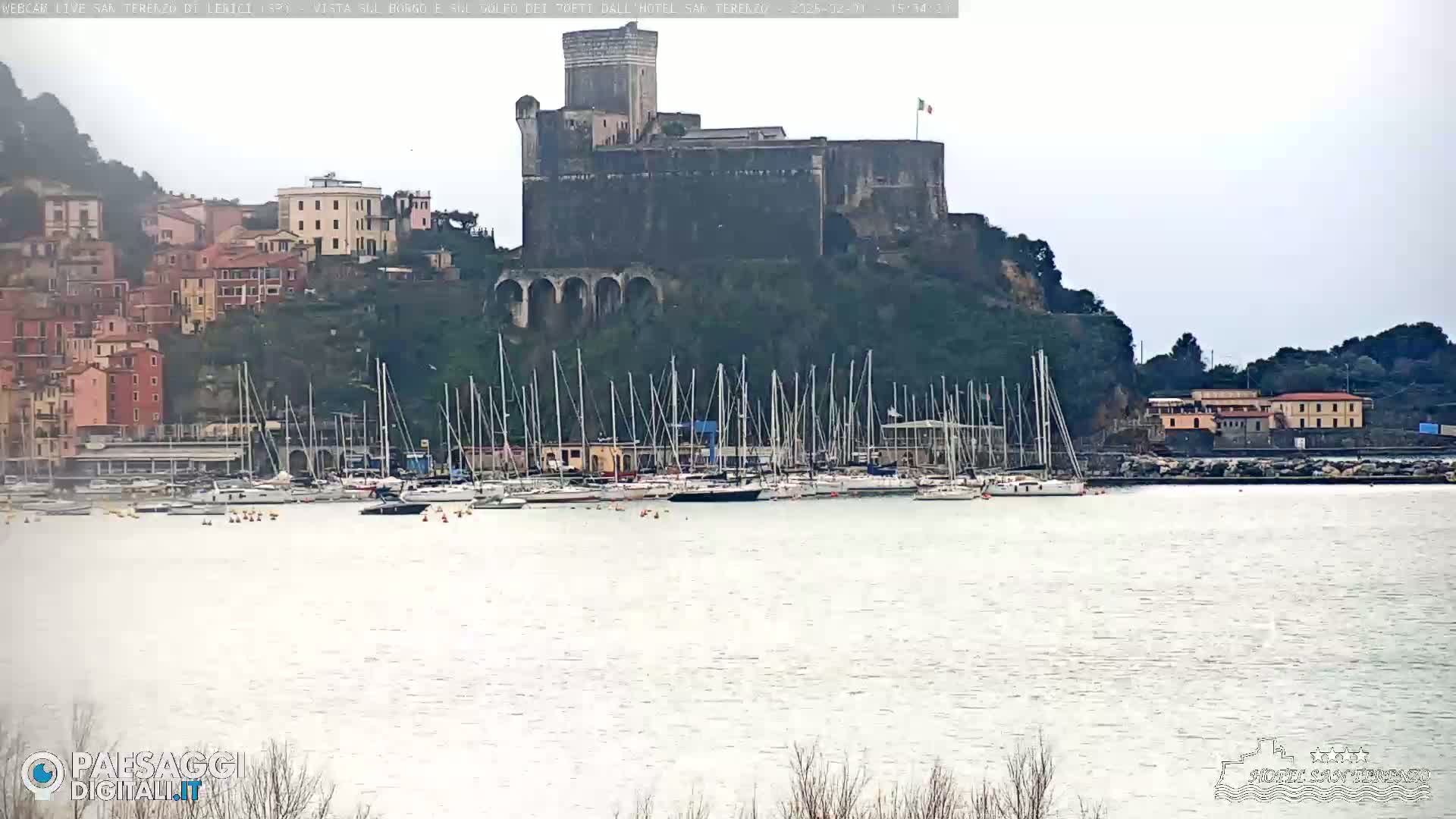 A stone castle sits atop a hill overlooking a harbor filled with sailboats on an overcast day.