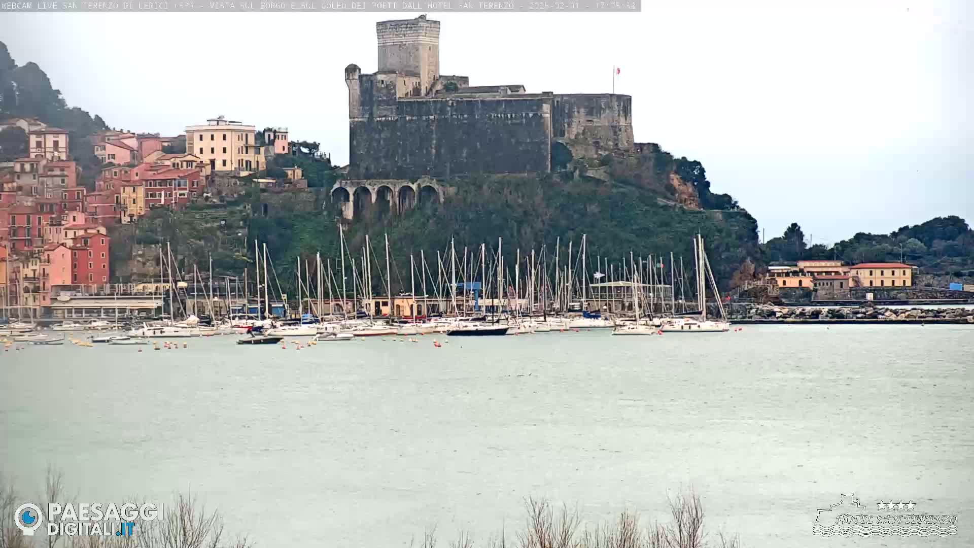 A stone castle overlooks a harbor filled with sailboats on an overcast day.