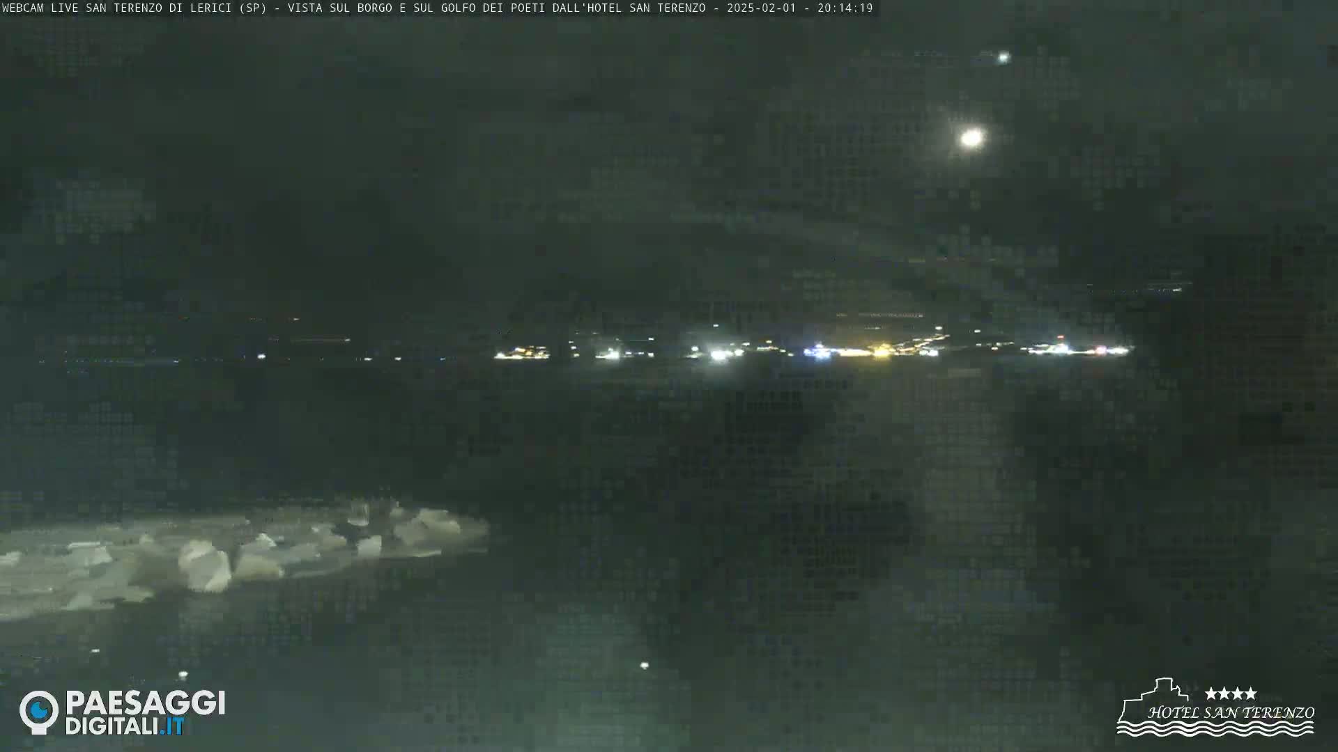 A nighttime view of a dark body of water with a faintly lit shoreline in the distance under a partly cloudy sky with a bright moon.