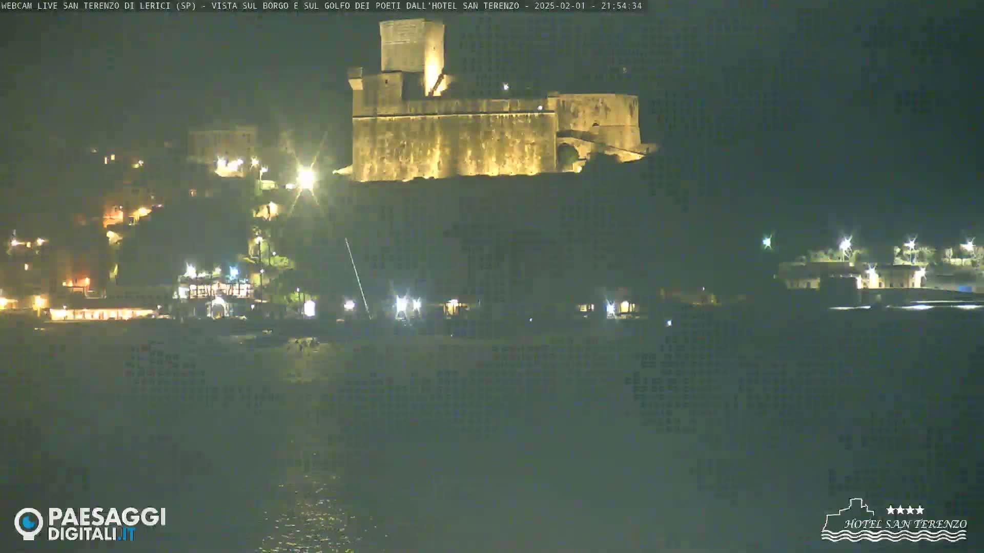 A nighttime view of a large, illuminated stone structure, possibly a castle, situated on a hill overlooking a coastal town with scattered lights.