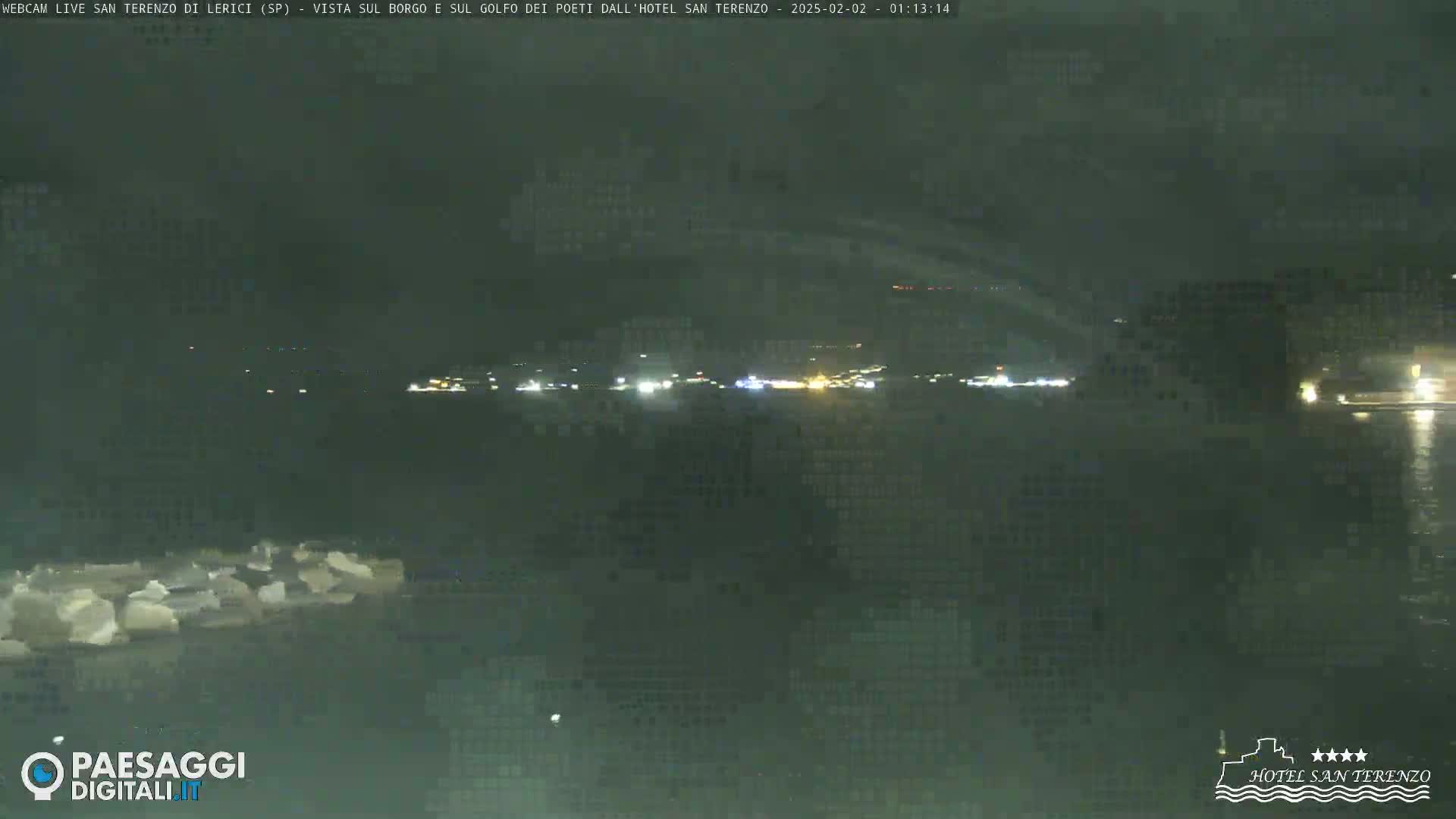 A nighttime view across a body of water shows a dark shoreline with faint lights in the distance under a cloudy sky.