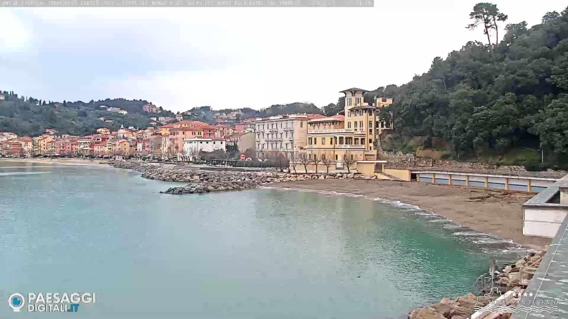 A seaside town with multi-story buildings sits along a calm bay under an overcast sky.