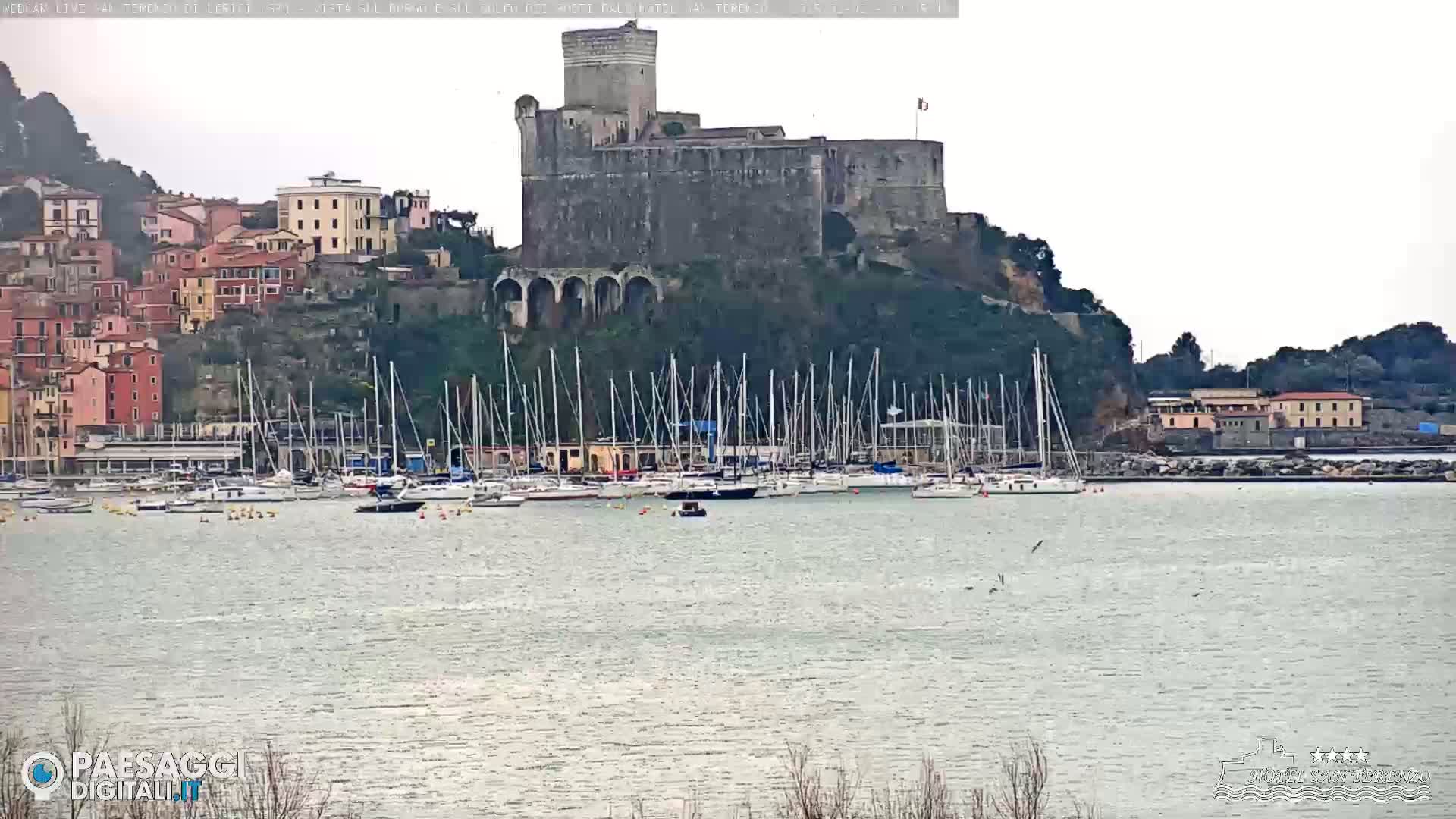 A stone castle sits atop a hill overlooking a harbor filled with sailboats, under an overcast sky.