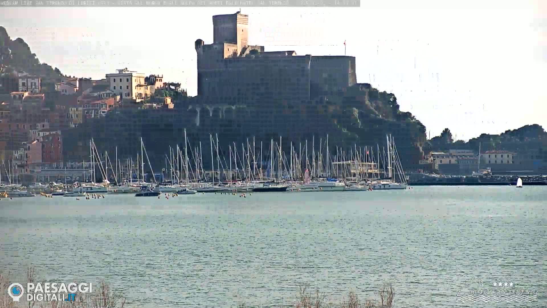A large castle sits atop a hill overlooking a harbor filled with sailboats on a calm, bright day.