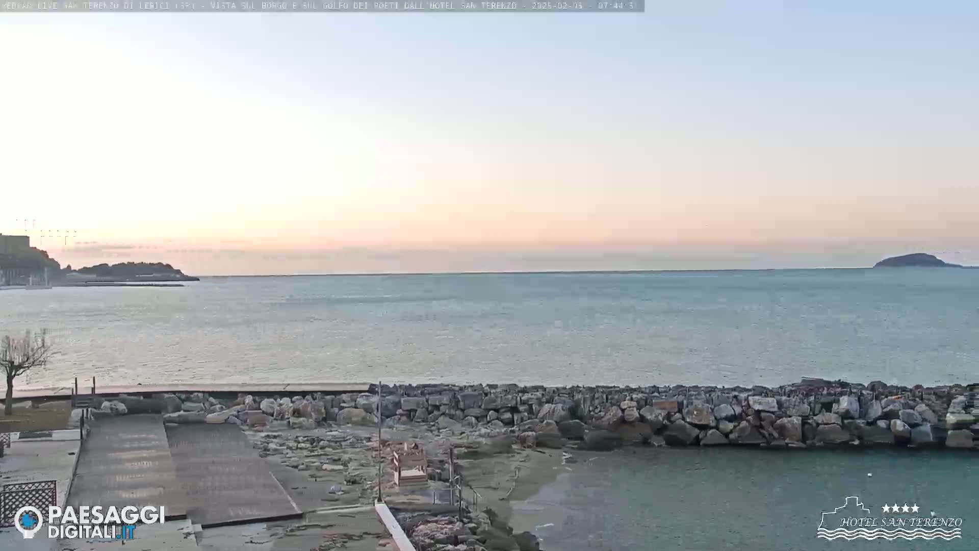 A calm sea at sunrise is viewed from a rocky shoreline, with a distant landmass visible on the horizon.
