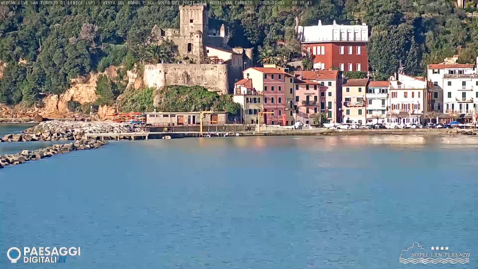 A calm body of water sits in front of a waterfront town, nestled against a hillside with a stone castle and colorful buildings under a sunny sky.
