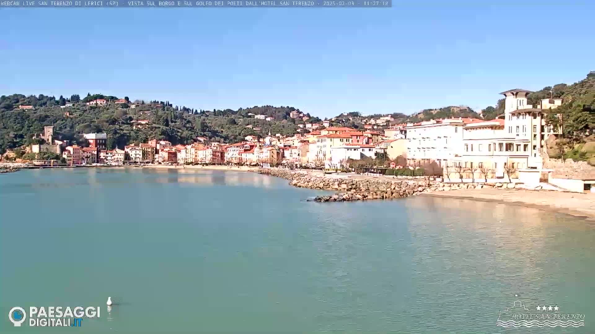 A coastal town with pastel-colored buildings sits on a hillside overlooking a calm, light-blue bay under a clear blue sky.