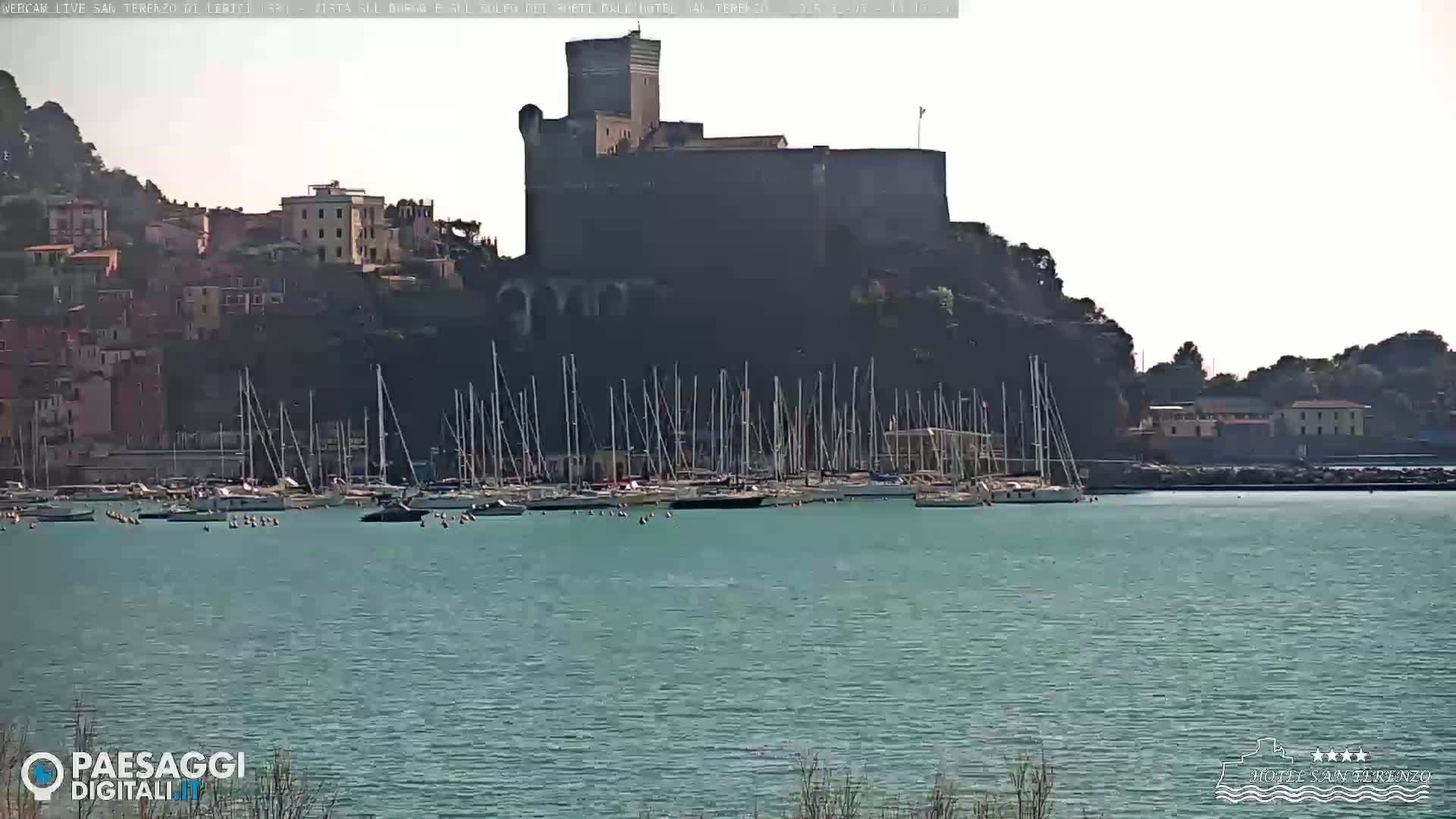 A large stone castle sits atop a hill overlooking a harbor filled with sailboats on a calm, sunny day.