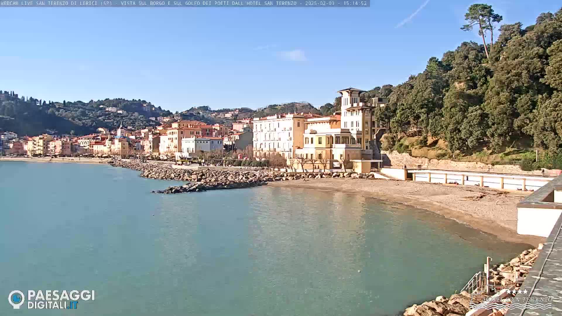 A coastal town nestled against a hillside, with a sandy beach and calm, blue-green water under a clear, sunny sky.
