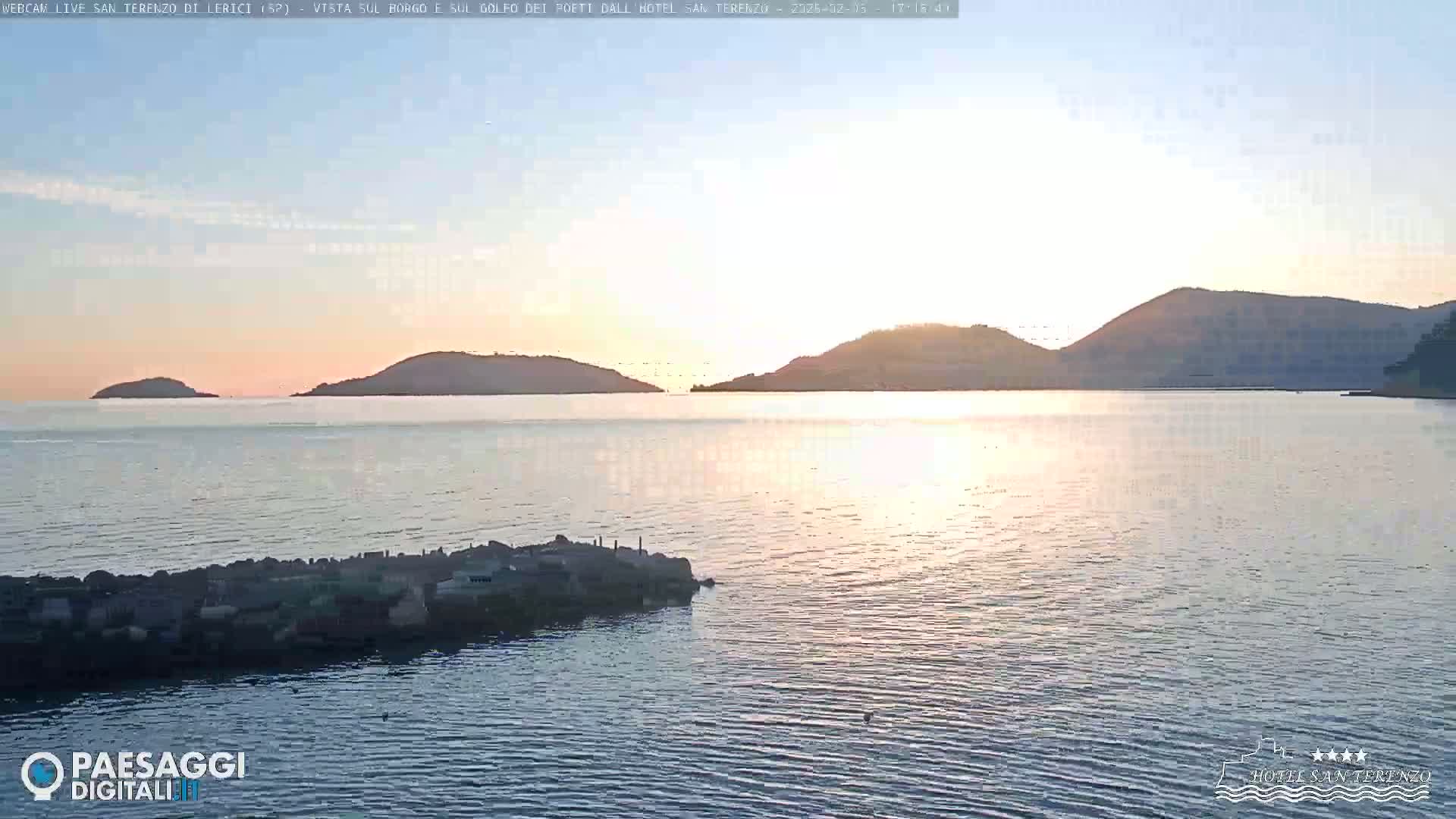 A calm body of water at sunset is seen, with several hills and a rocky jetty in the foreground.