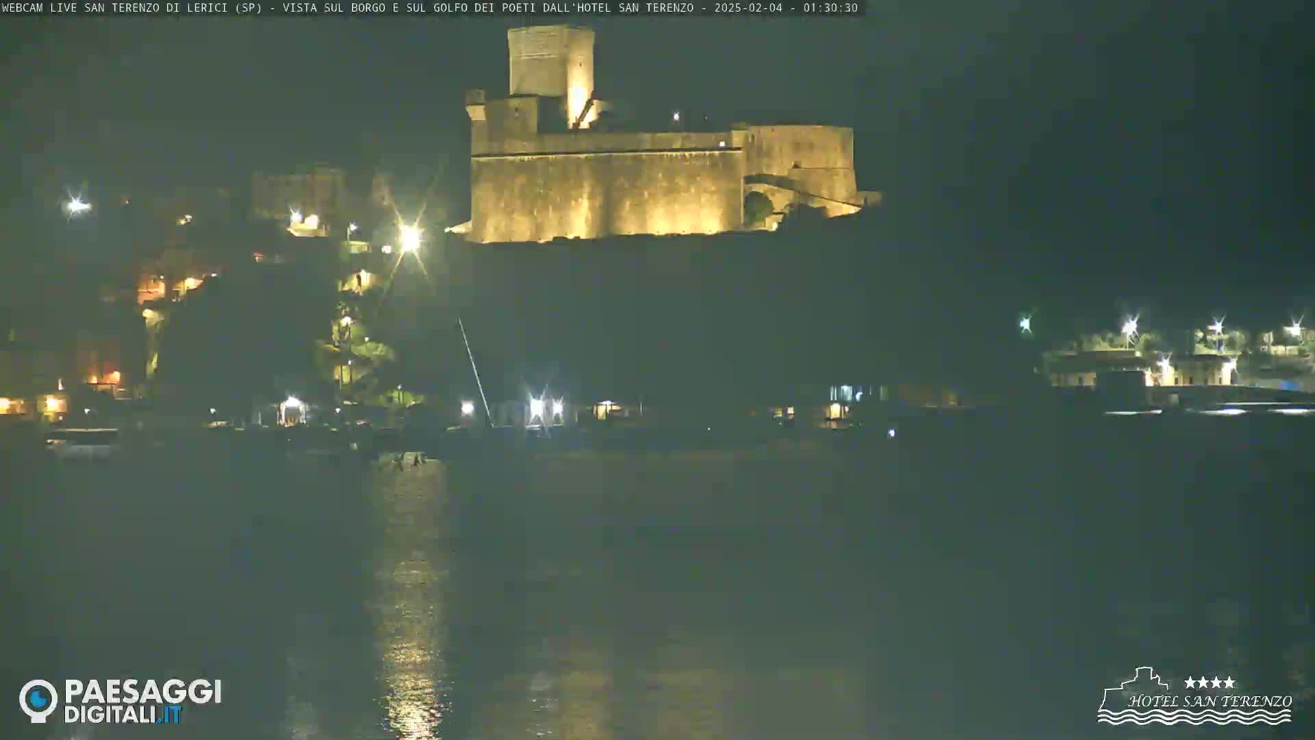 A dark, nighttime view of a lit-up stone castle perched atop a hill overlooking a calm body of water.