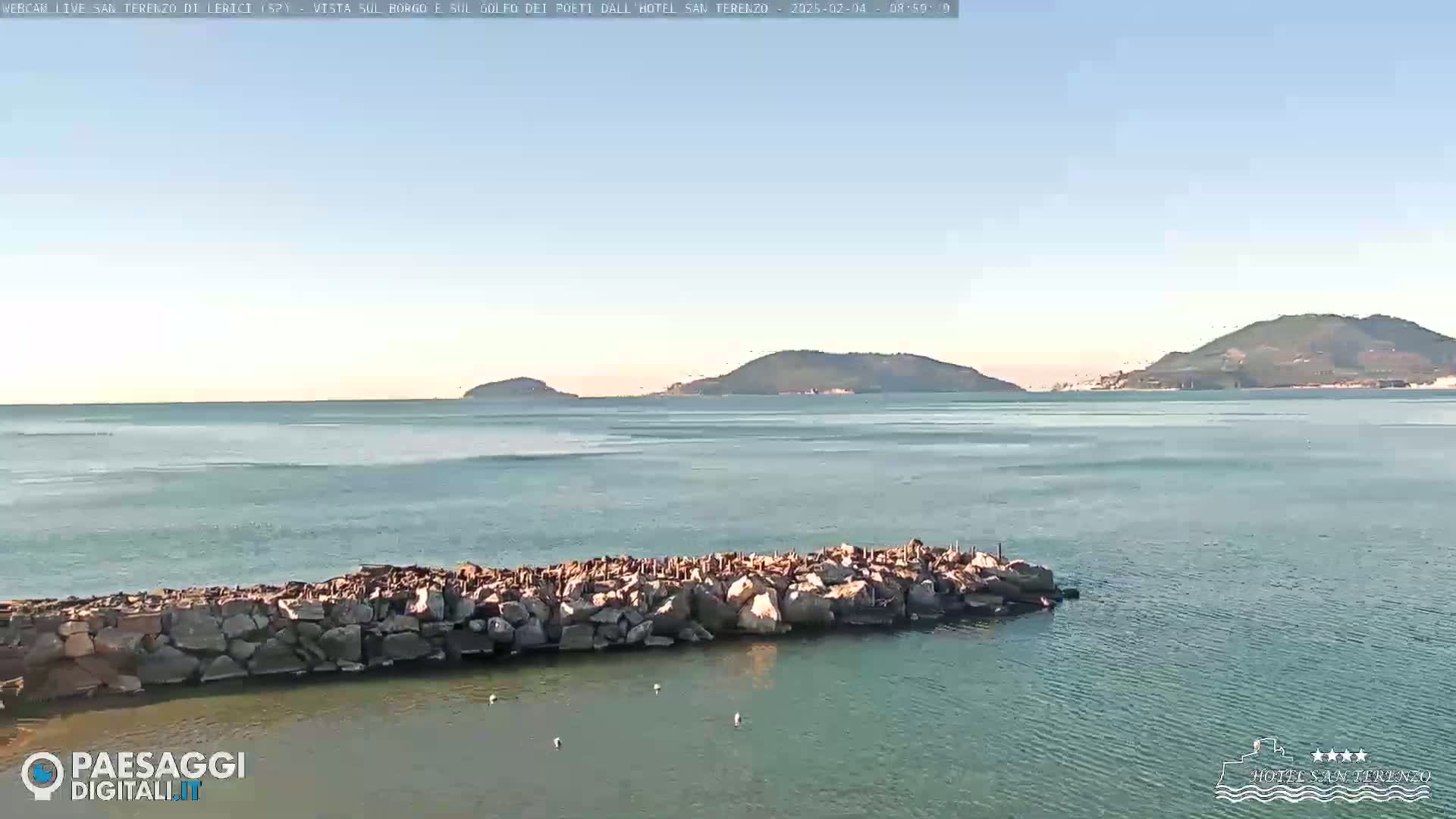 A calm sea with a rocky breakwater in the foreground and distant hills under a clear sky.
