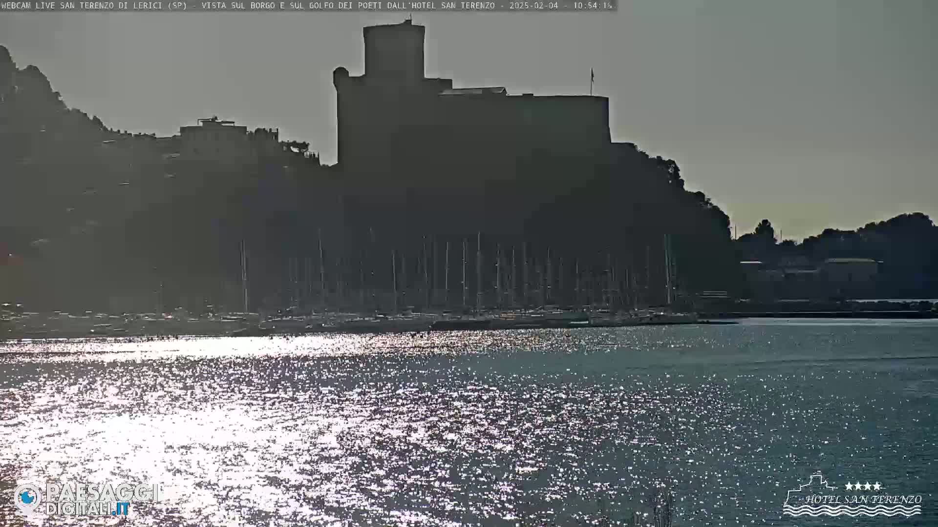A dark castle sits atop a hill overlooking a harbor filled with sailboats, under a bright, mostly clear sky reflecting on the water.