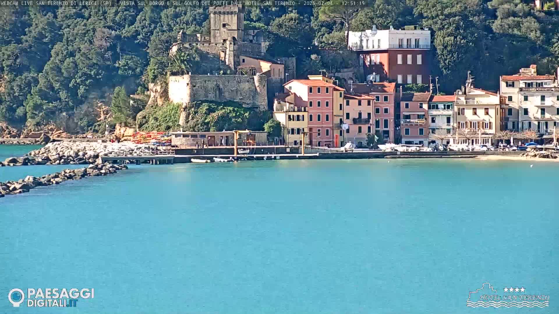 A coastal town with colorful buildings, a stone fortress, and a small harbor sits on a hillside overlooking a calm, turquoise sea under a sunny sky.