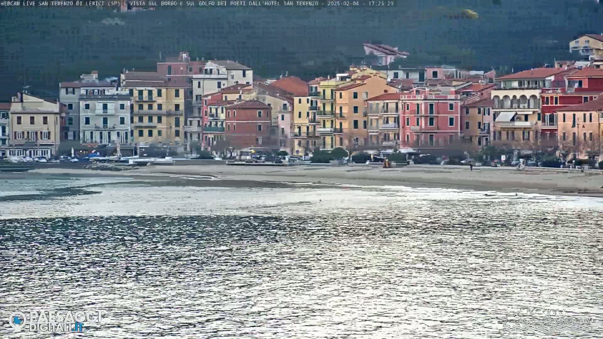 A row of colorful buildings lines a beach next to calm, slightly rippled water under a cloudy sky.