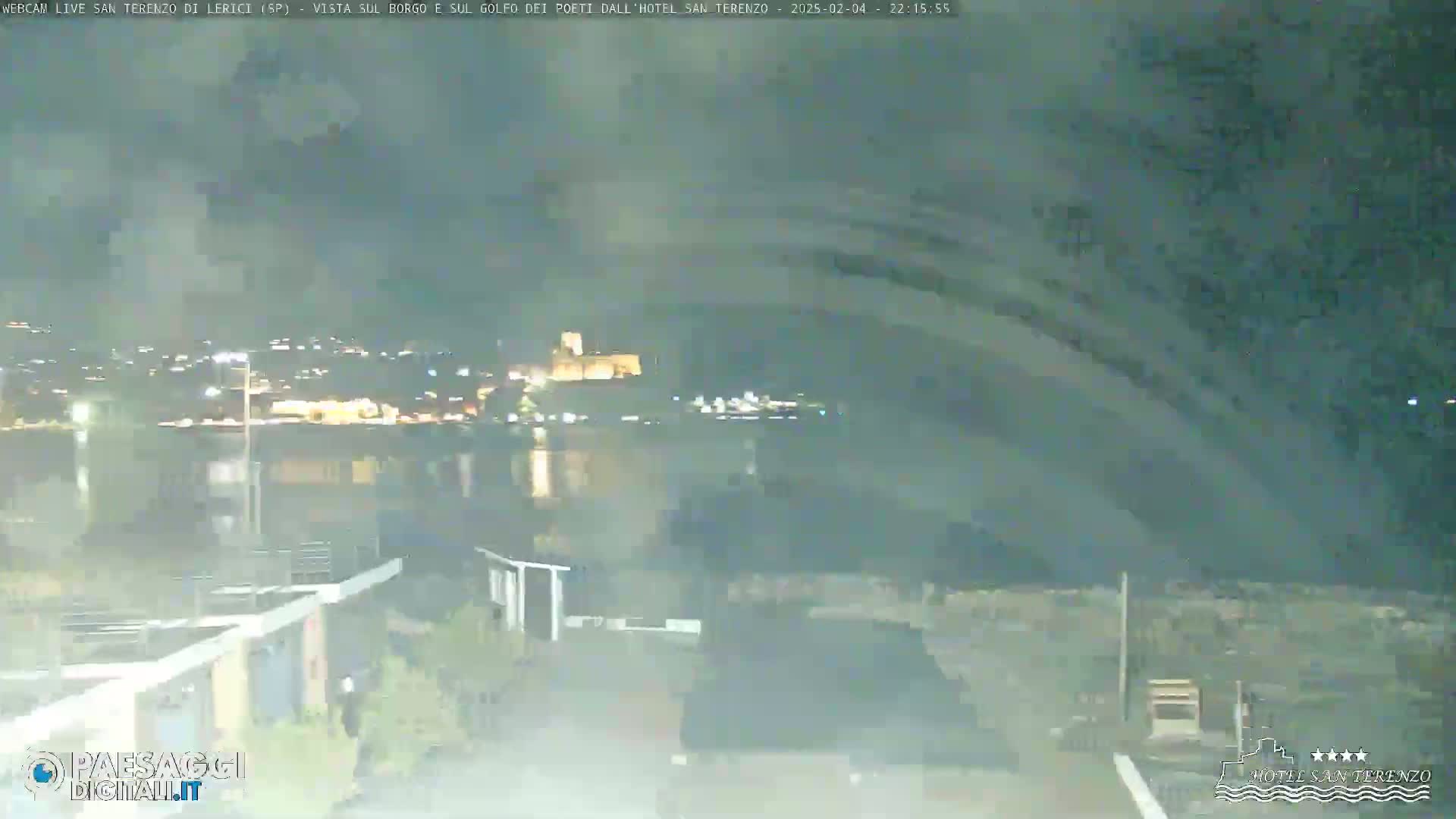 A nighttime, hazy view of a town across a body of water, with a castle visible in the distance.