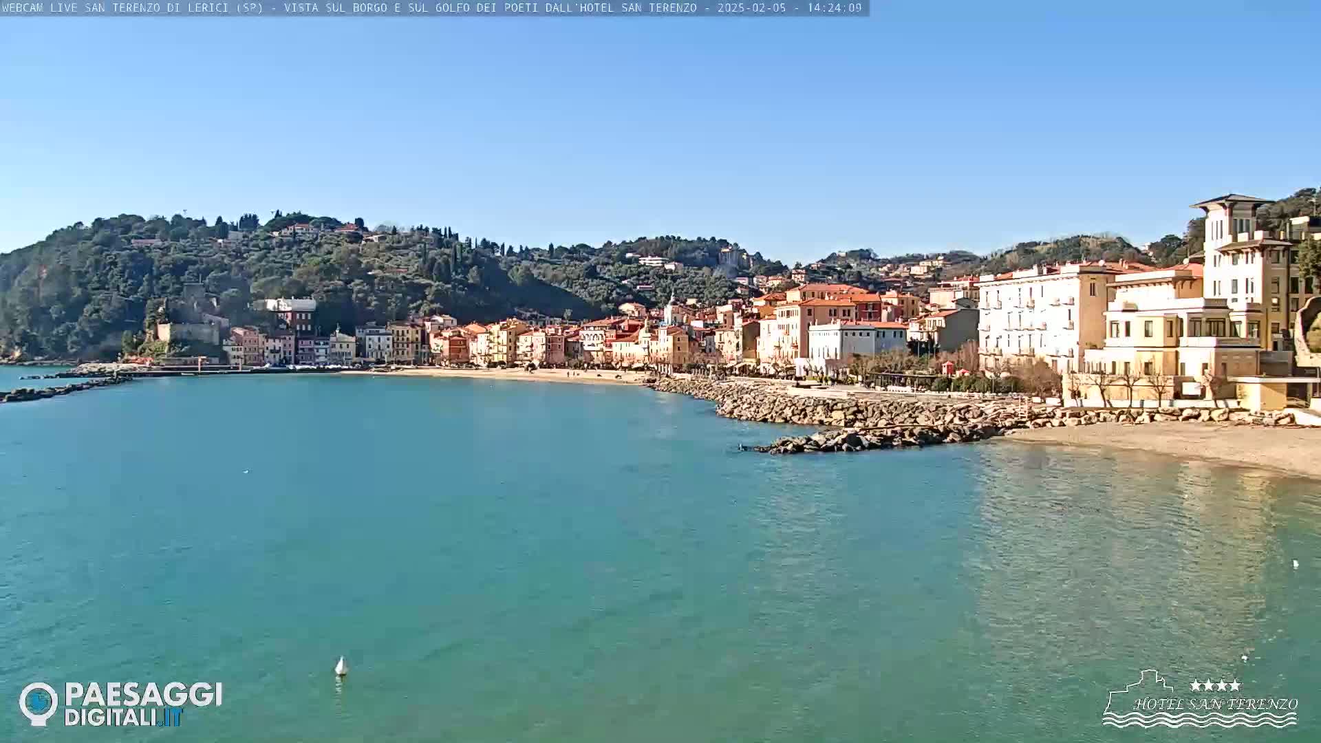 A picturesque coastal town with colorful buildings sits on a hillside overlooking a calm, turquoise bay under a clear blue sky.