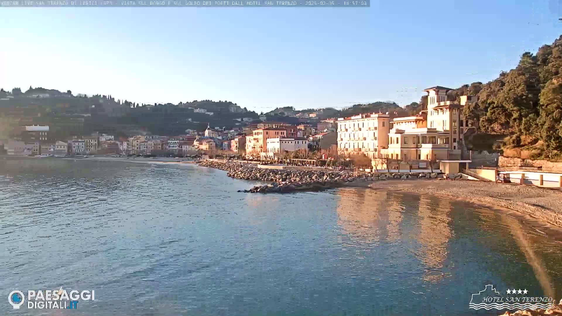 A calm, sunlit body of water laps a pebbled beach in front of a coastal town nestled against tree-covered hills.