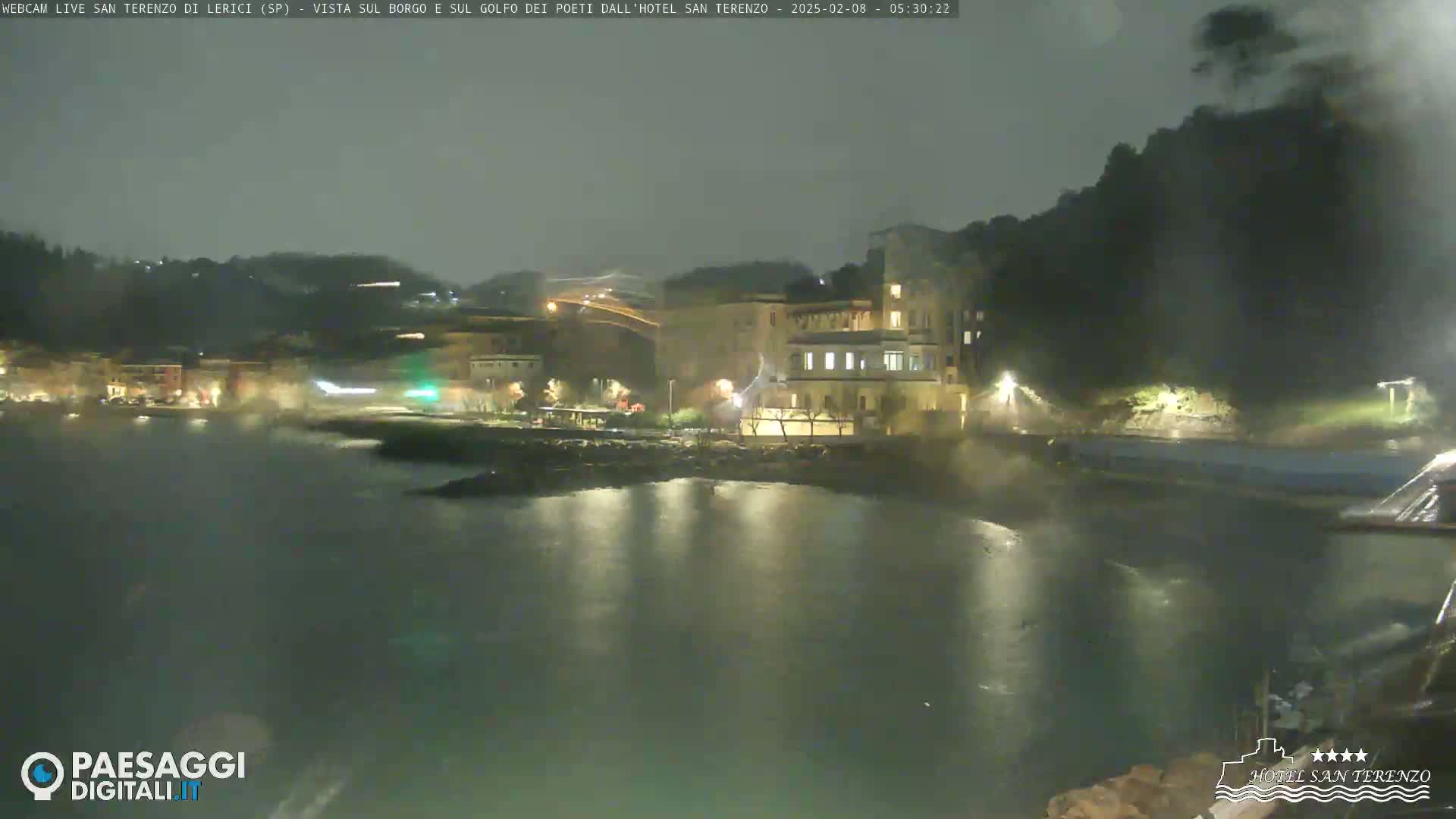A nighttime view of a coastal town with buildings illuminated near a dark body of water under an overcast sky.