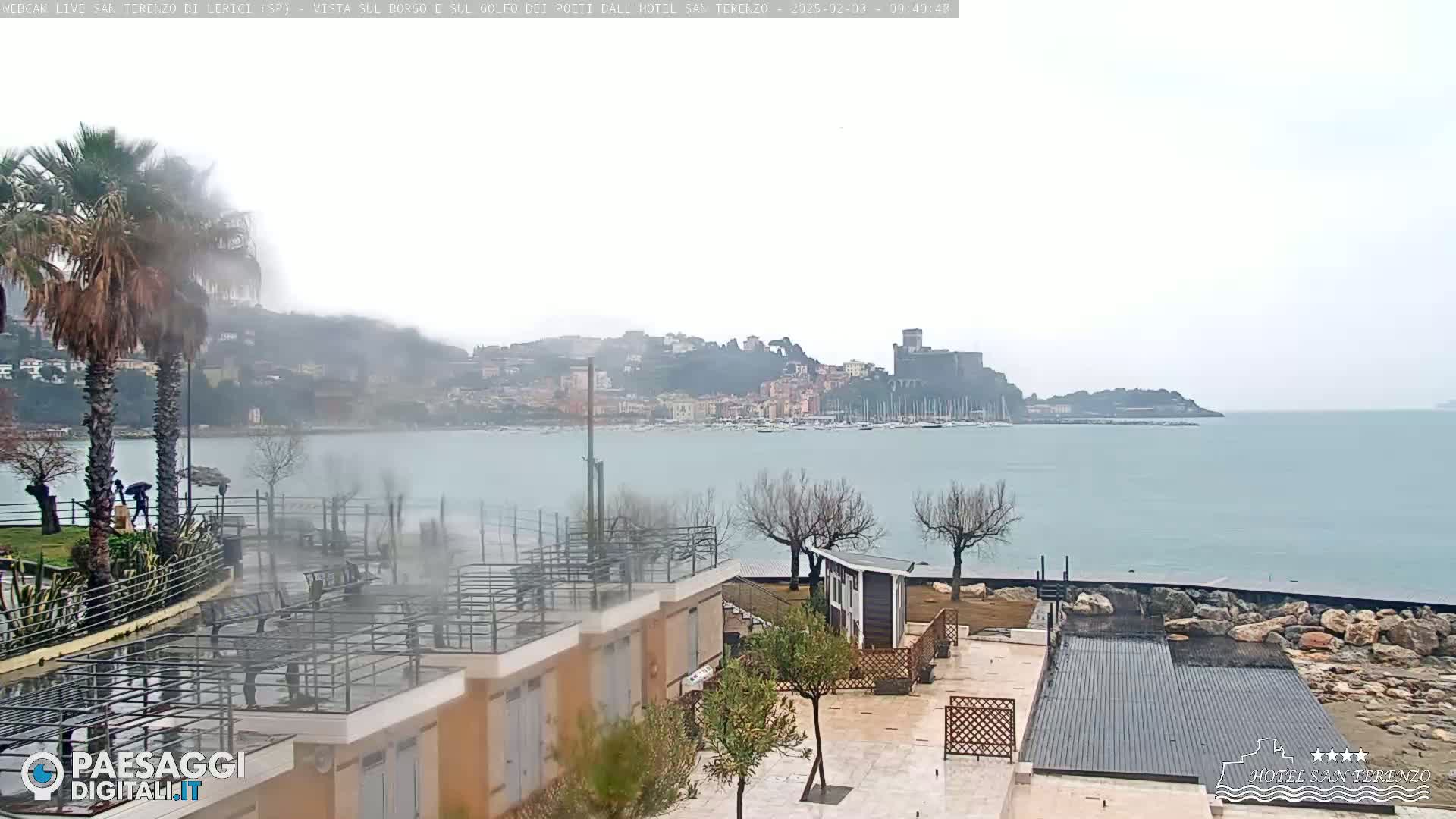 A coastal town with a castle is visible across a calm body of water on an overcast day, with palm trees and modern buildings in the foreground.
