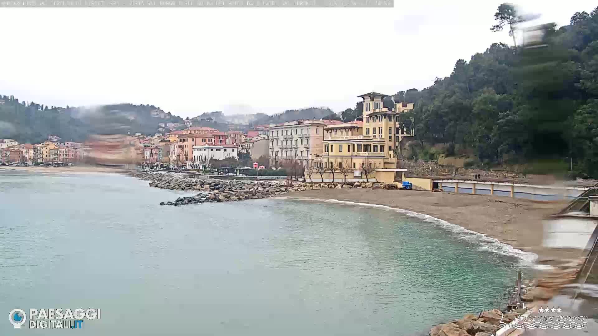 A coastal town with multi-story buildings sits on a beach next to calm, greenish-blue water under an overcast sky.