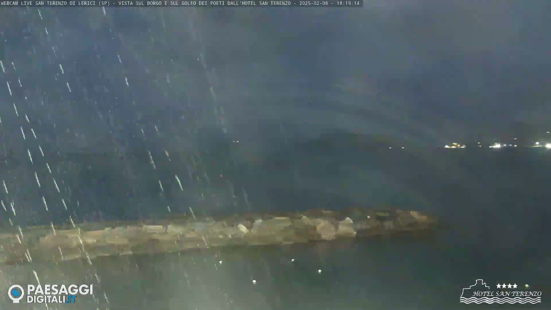 A nighttime view of a rocky shoreline under heavy rain, with lights faintly visible across the water in the distance.