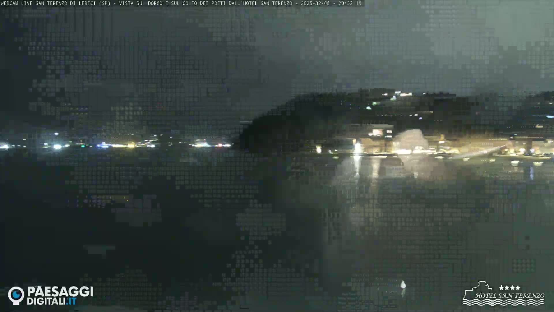 A nighttime view of a dark body of water reflecting lights from a town on the opposite shore under a cloudy sky.