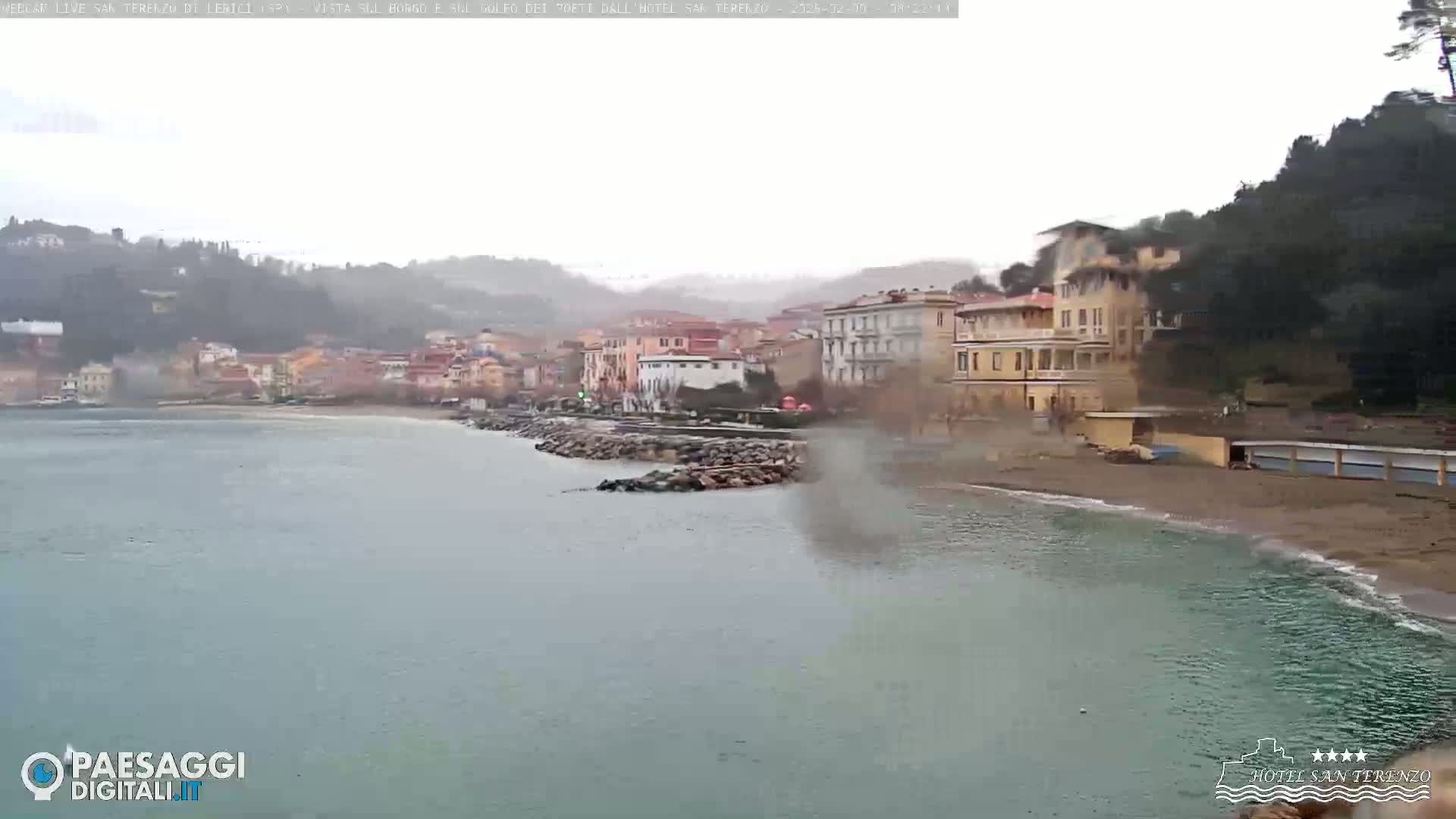 A coastal town is seen on an overcast day with a calm sea; the town is nestled between hills, and a sandy beach is visible in the foreground.