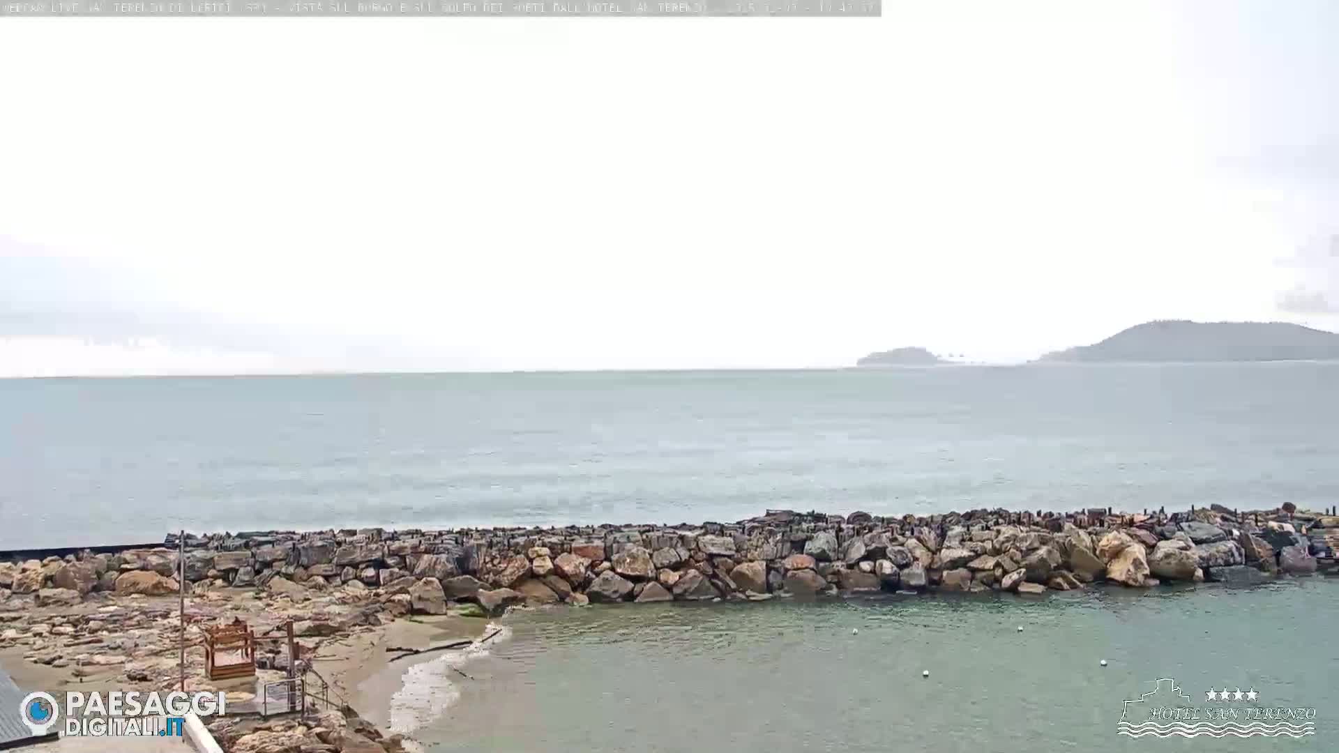 A rocky breakwater protects a calm, shallow inlet from a choppy sea under an overcast sky, with distant hills visible on the horizon.