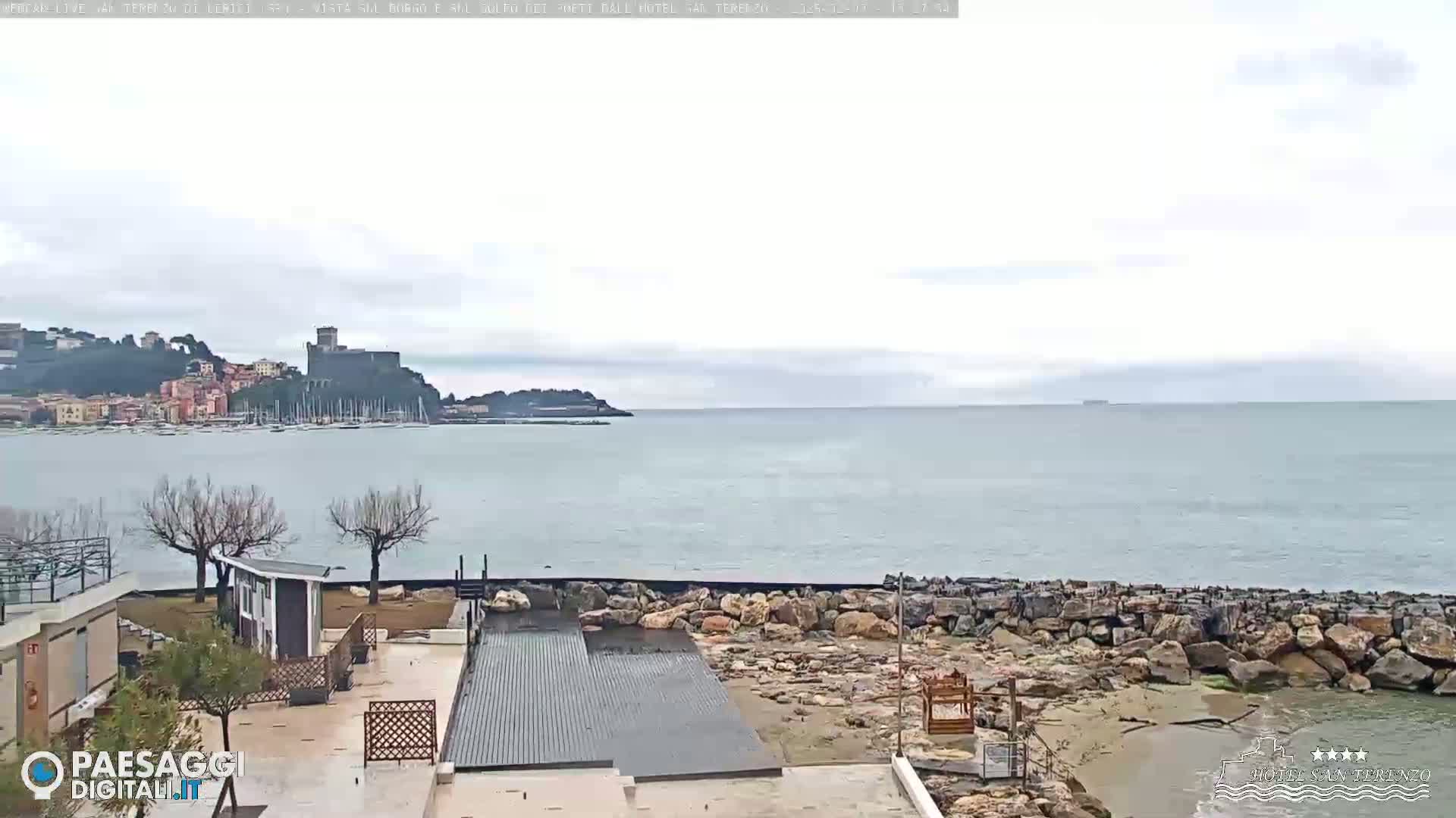 A seaside view shows a calm sea under an overcast sky, with a town and castle visible in the distance, and a rocky shoreline in the foreground.