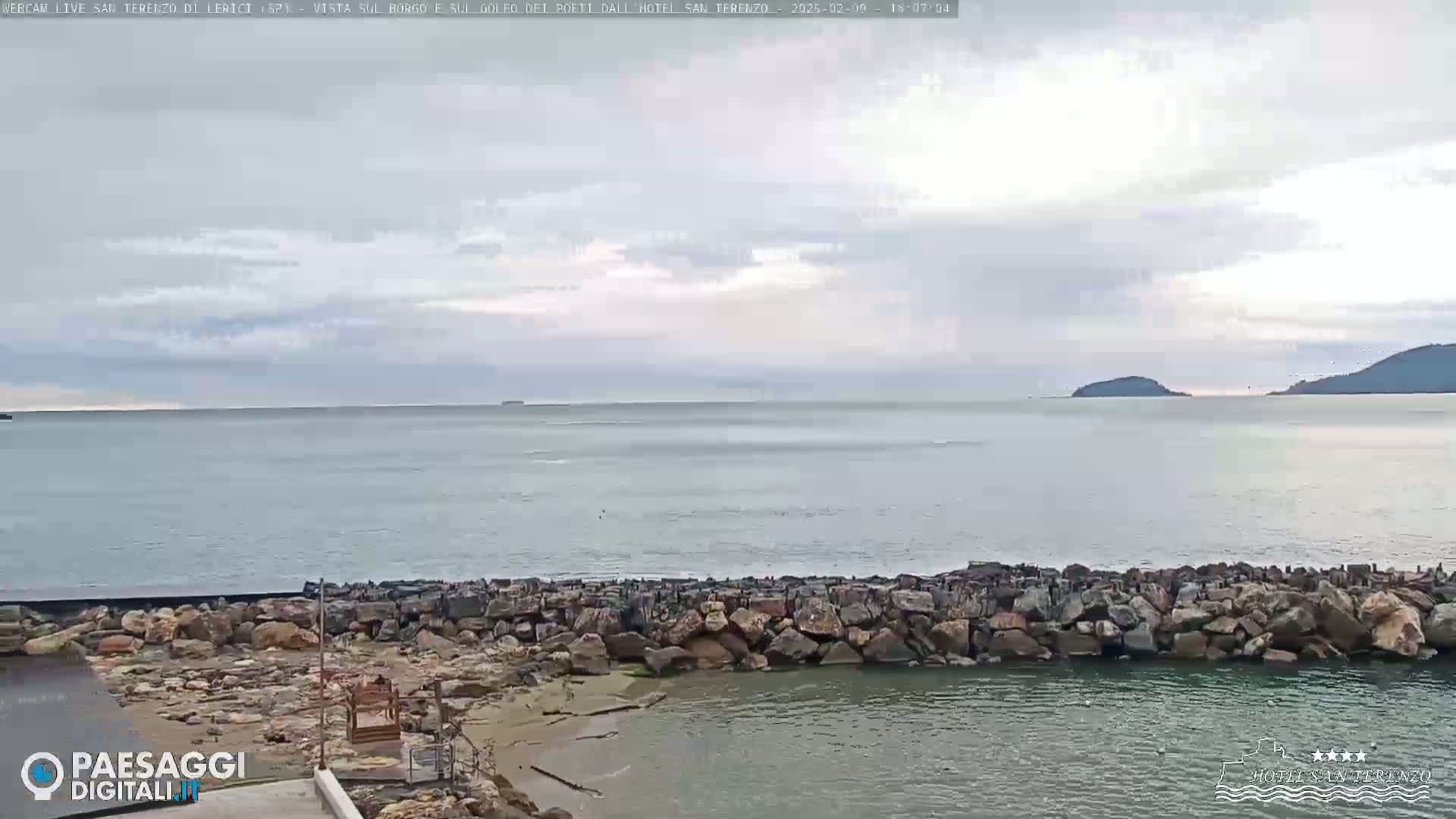 A rocky seawall borders a calm sea under a cloudy sky, with distant islands visible on the horizon.
