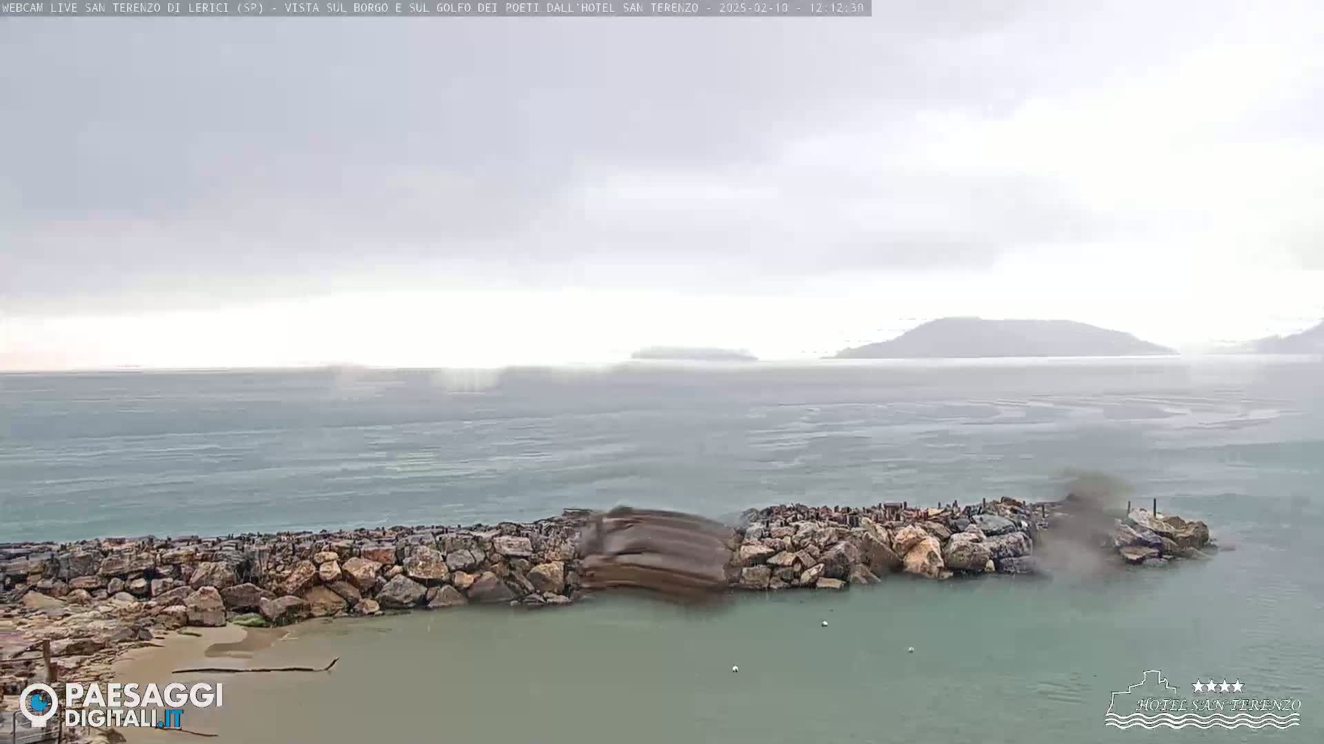 A rocky breakwater juts into a choppy sea under an overcast sky, with distant hills barely visible in the hazy distance.