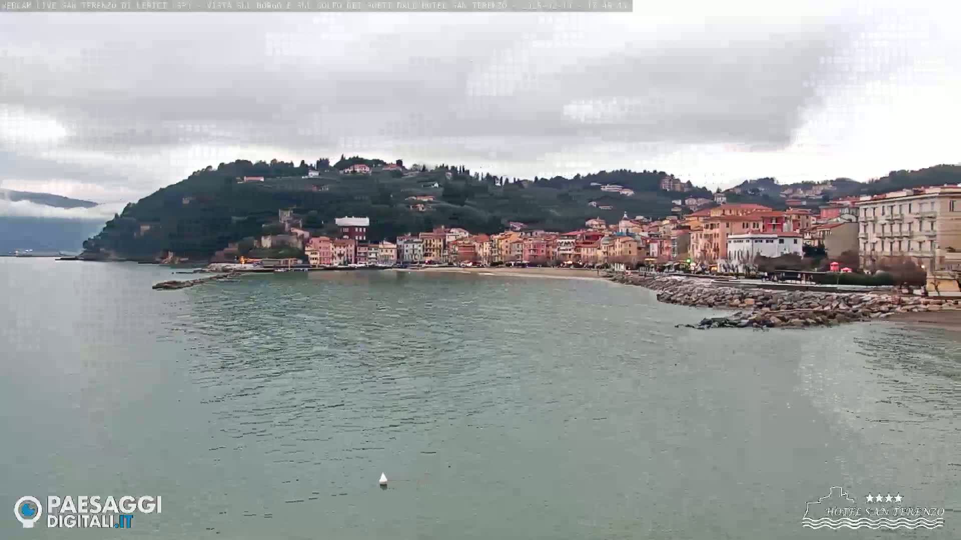 A coastal town with multi-story buildings is nestled against a hillside under a cloudy sky, with calm, grey-green water in the foreground.