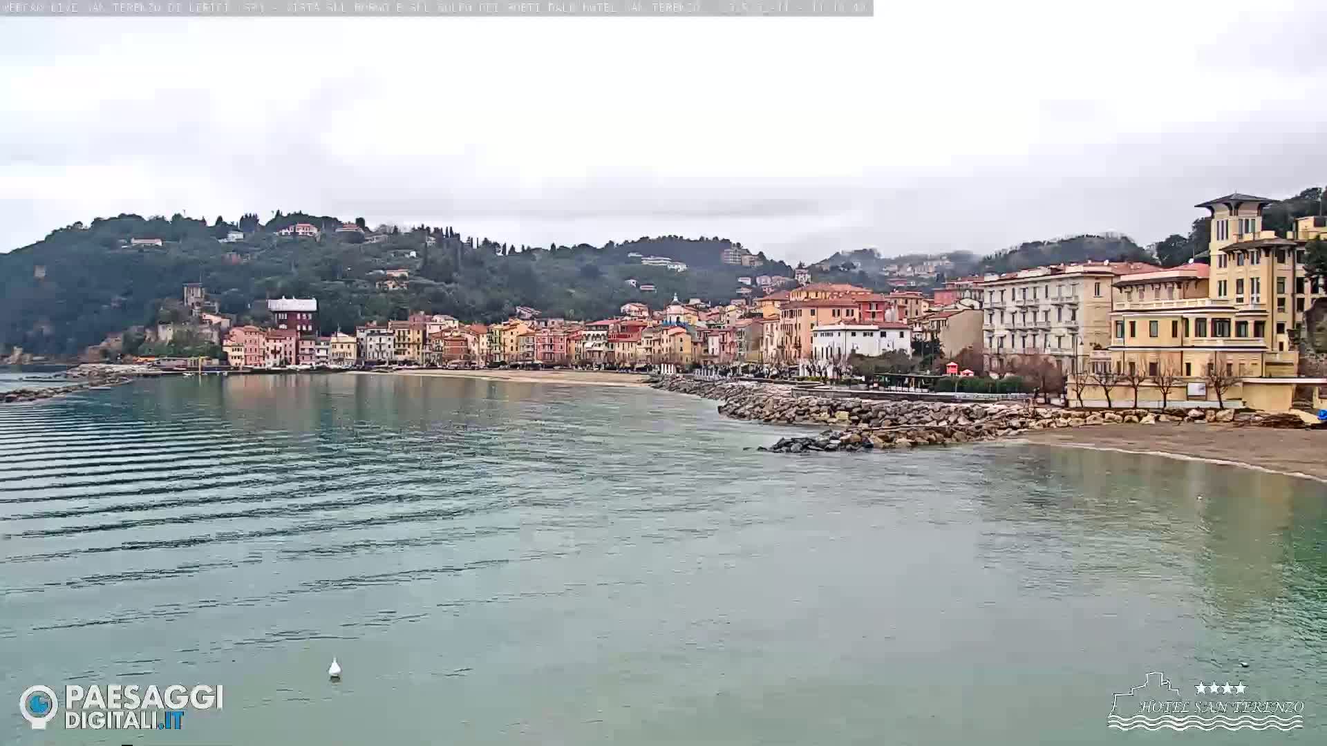 A coastal town nestled against a hillside, with calm, slightly rippled water in the foreground under an overcast sky.