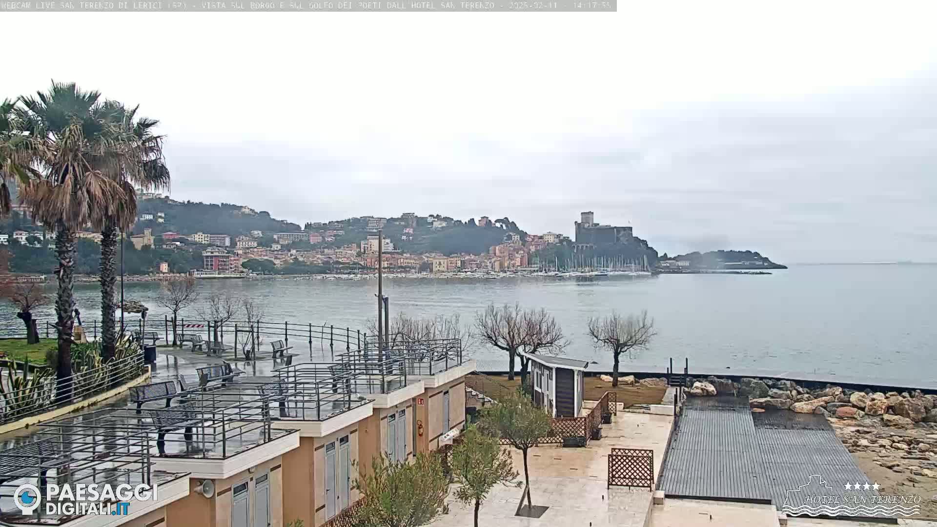 A seaside town with a castle is visible across a calm body of water under an overcast sky, seen from a waterfront area with buildings and landscaping.