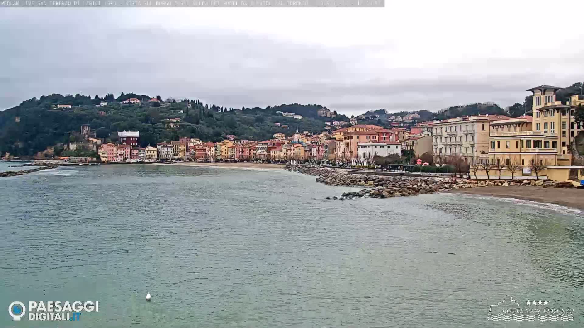 A coastal town with multi-story buildings sits on a hillside overlooking a calm, gray sea under an overcast sky.
