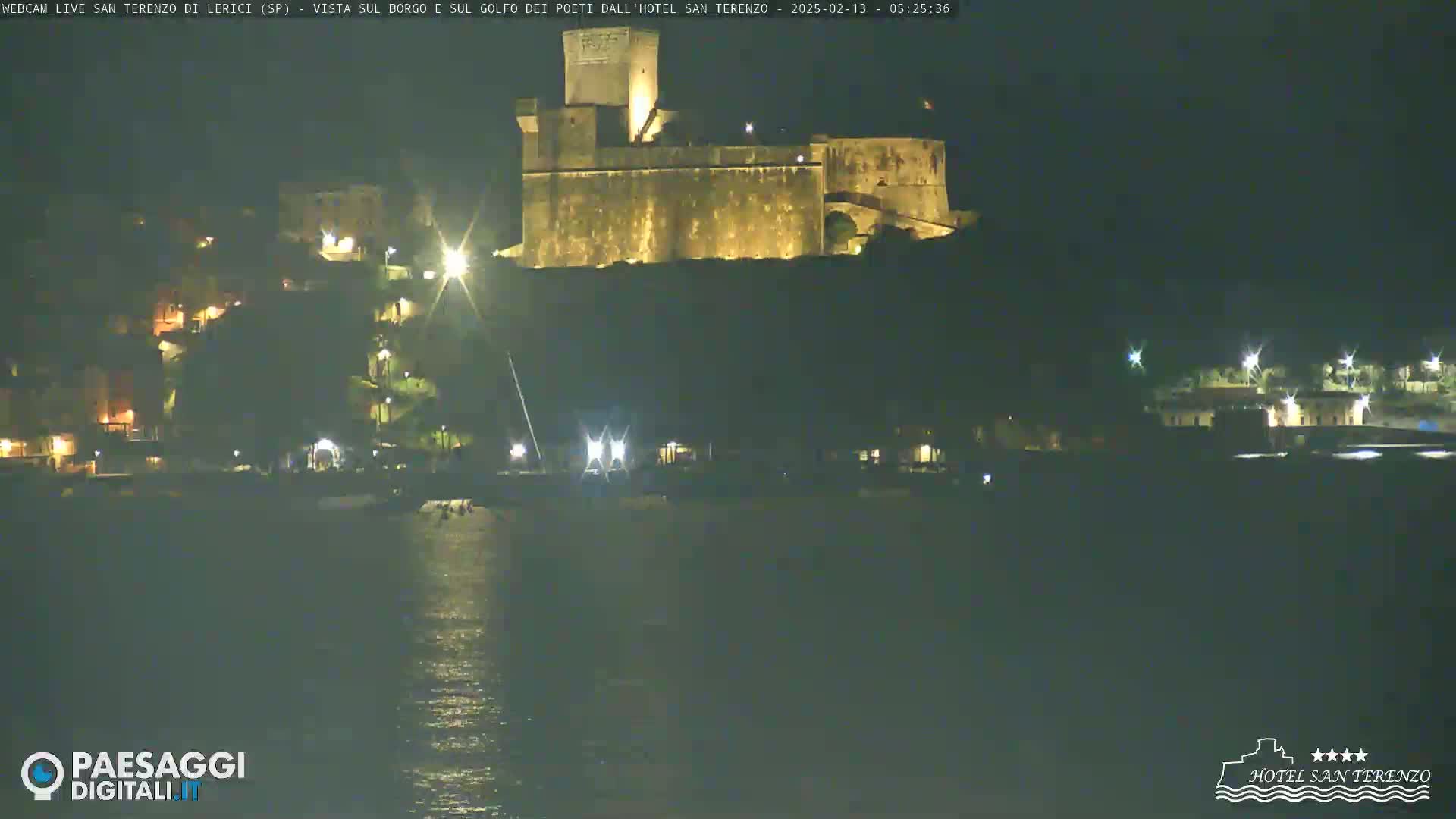 A nighttime view of a stone fortress illuminated on a hill overlooking a dark body of water and a sparsely lit shoreline.