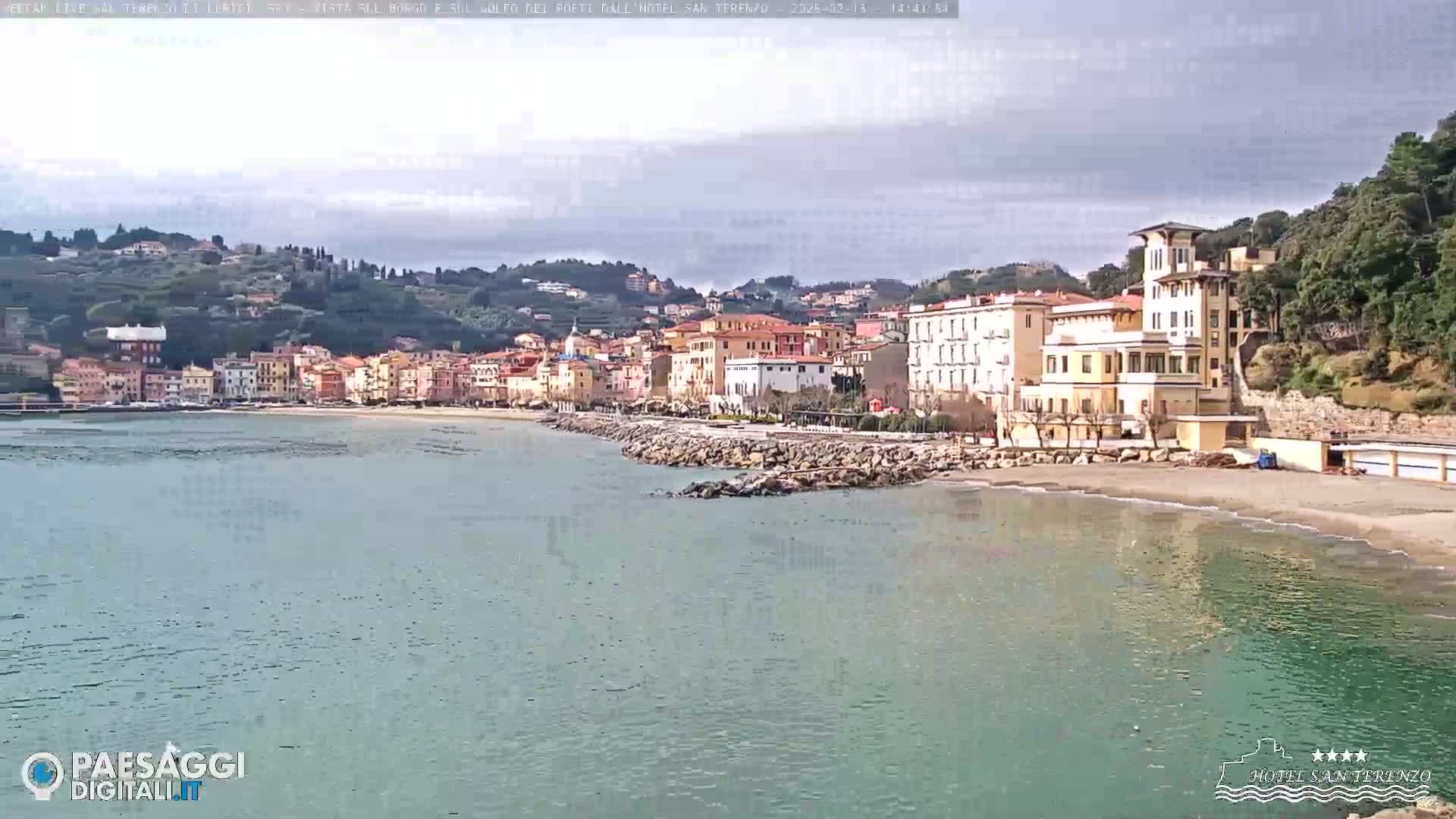 A coastal town with multi-story buildings sits on a gently sloping hill overlooking a calm sea under a mostly cloudy sky.