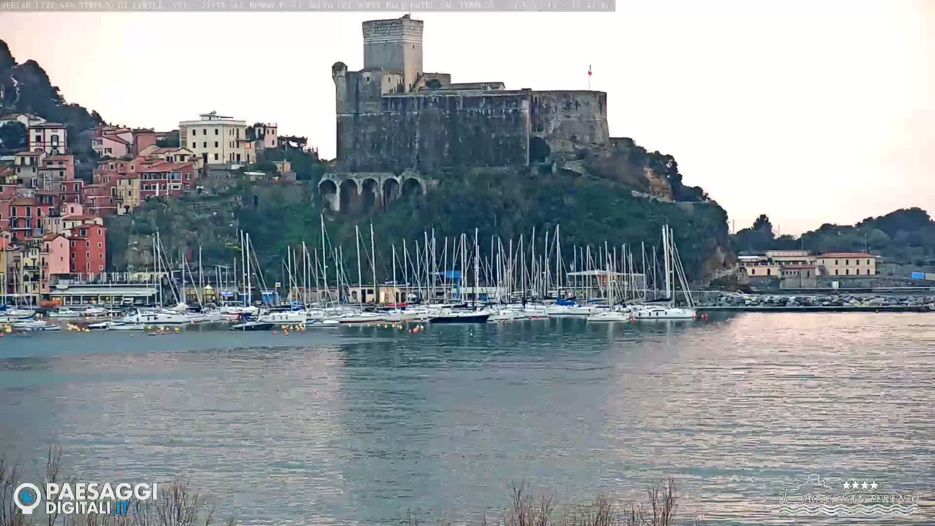 A stone castle sits atop a hill overlooking a harbor filled with sailboats, under a mostly clear, pale sky.