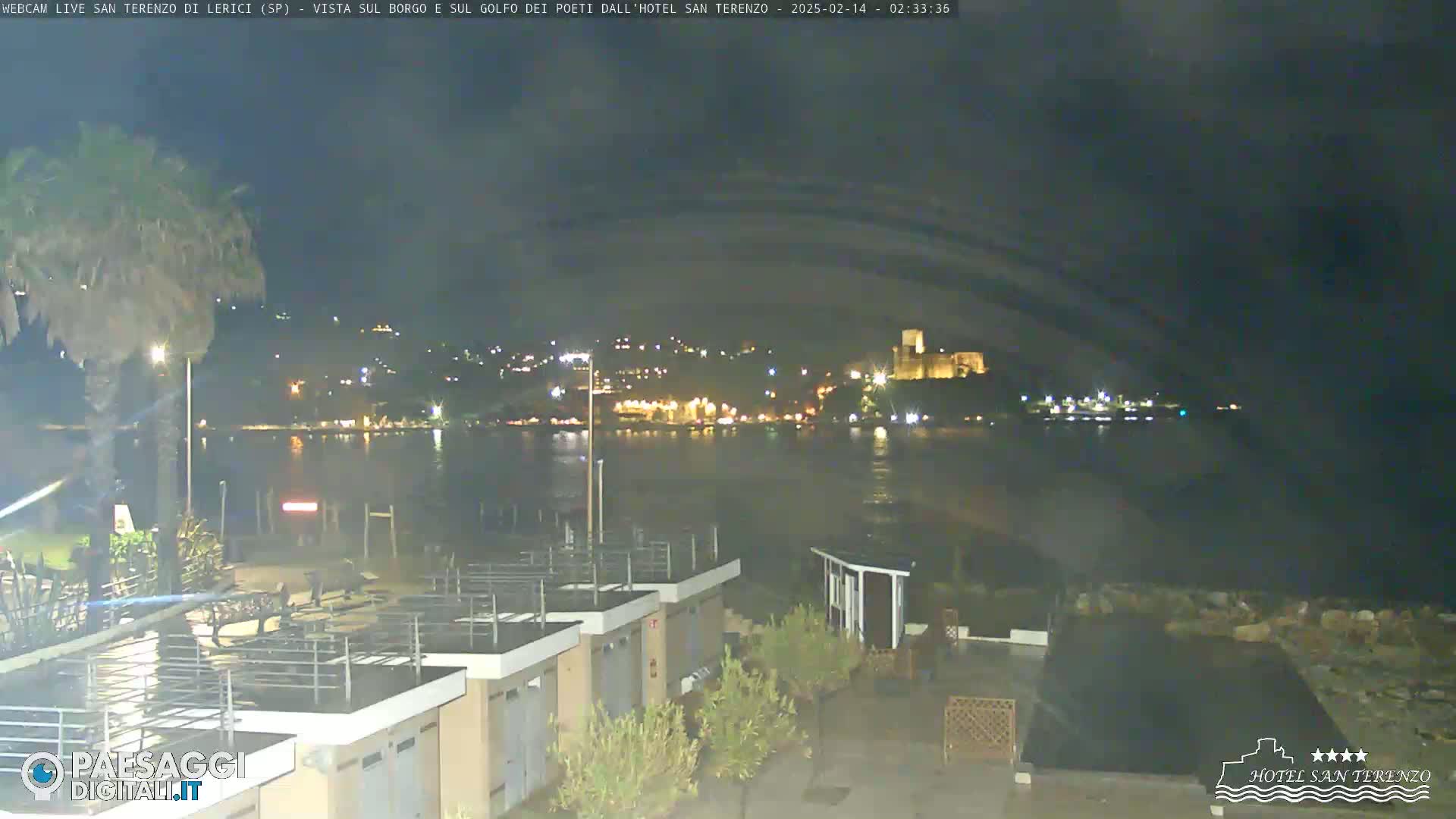 A nighttime seaside view shows a calm body of water reflecting a town's lights, with a castle visible in the distance, and beachside structures in the foreground under a mostly clear, dark sky.