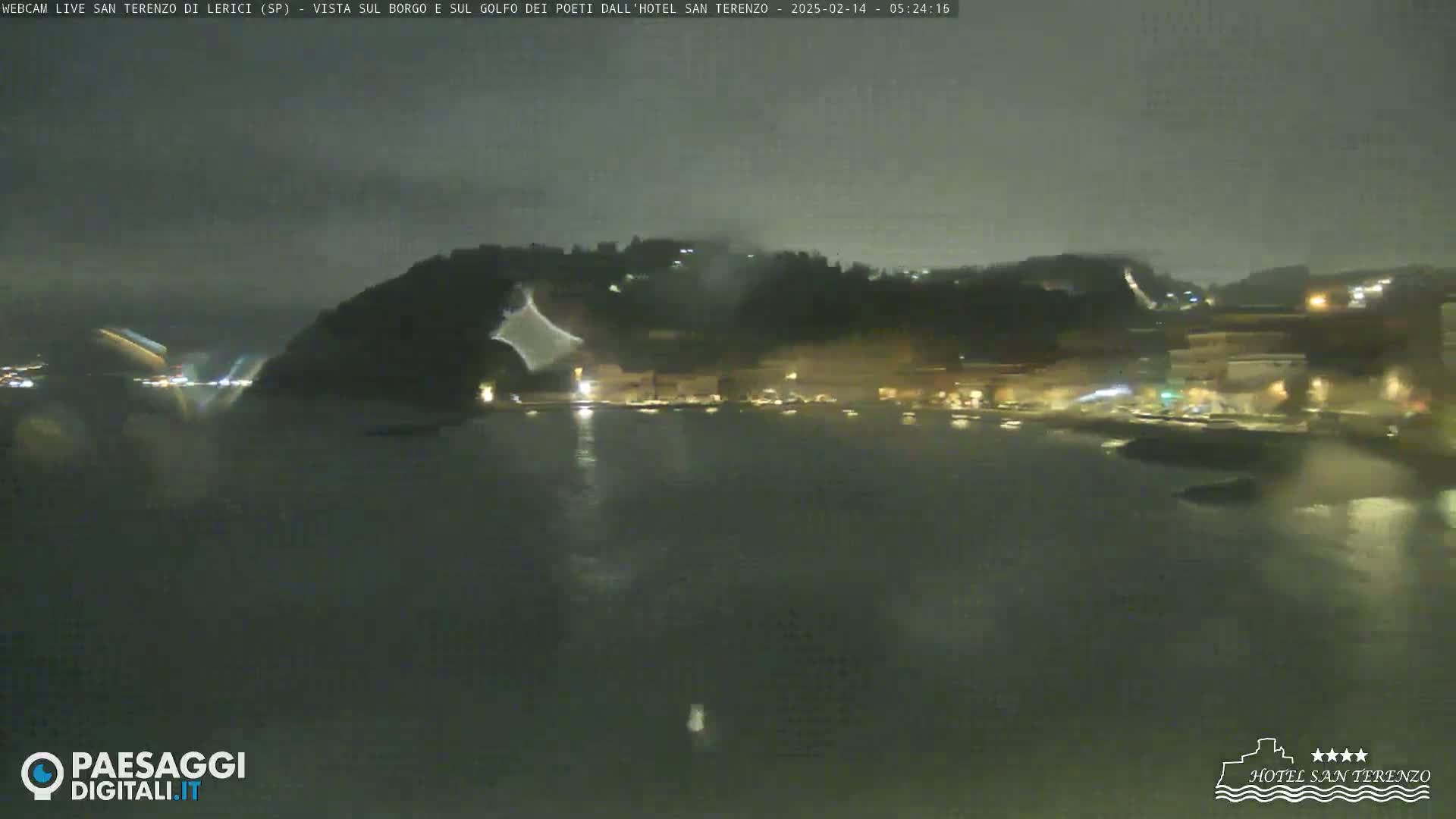 A nighttime, slightly rainy view of a calm body of water reflecting the lights of a town nestled against a dark hillside.