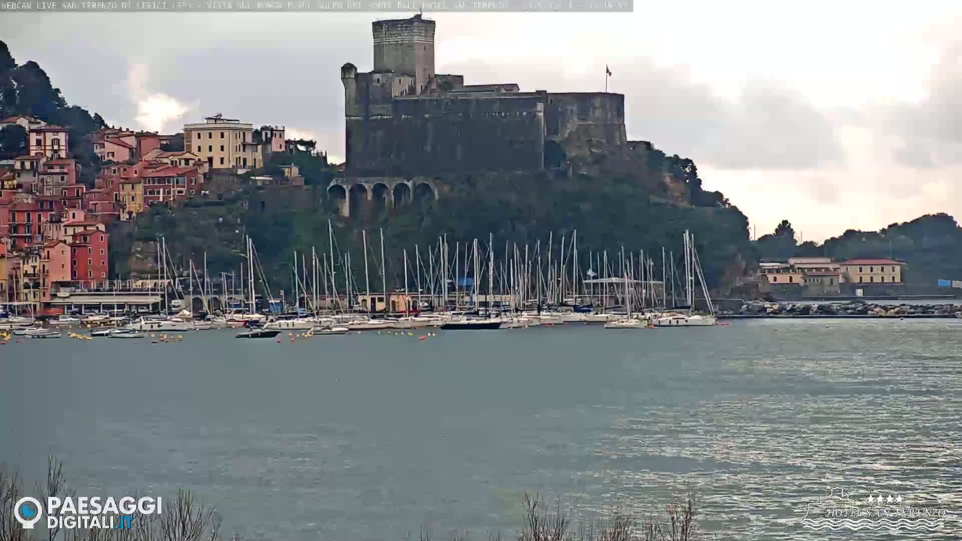 A stone castle overlooks a harbor filled with sailboats on an overcast day.